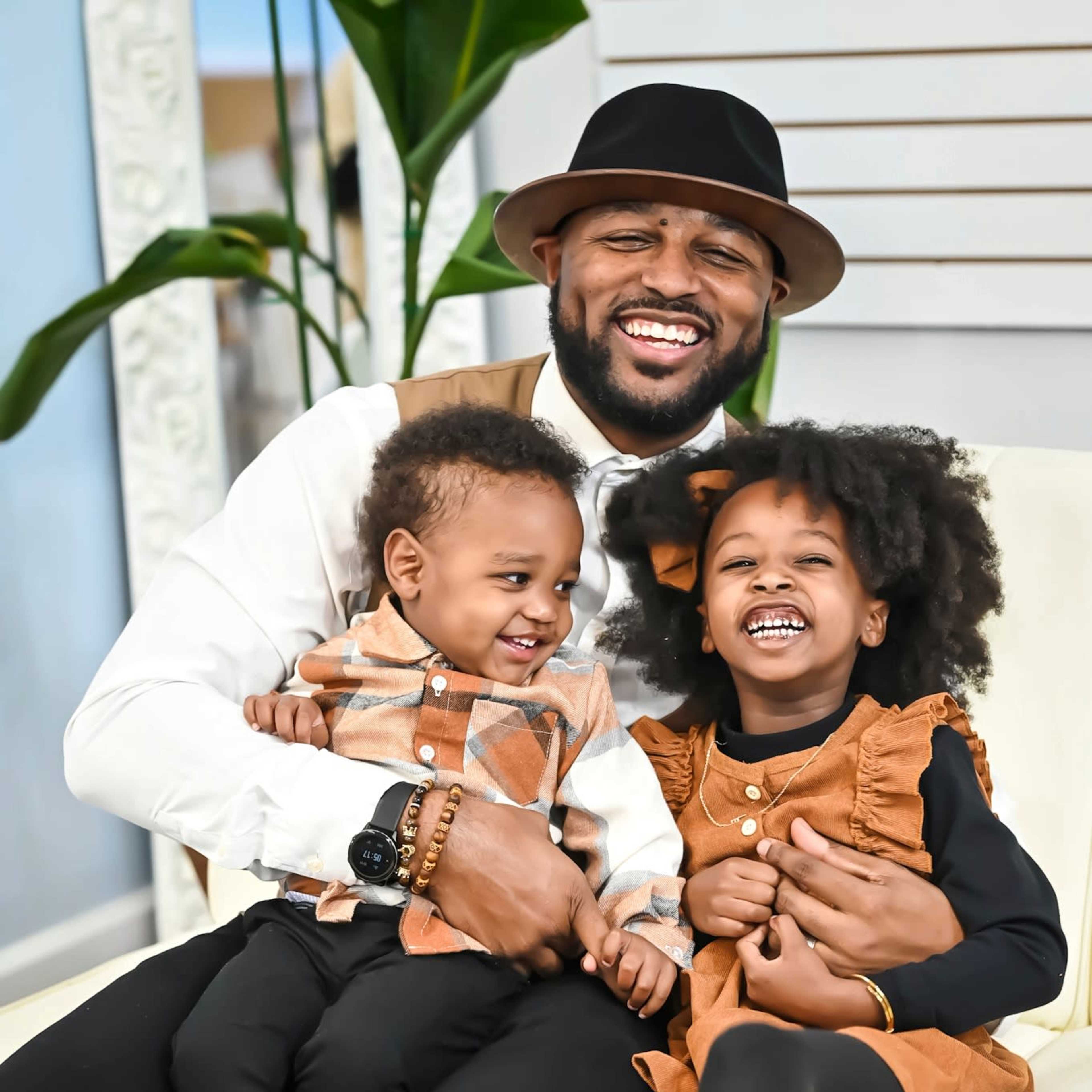 A man in formal attire sits on a white chair, joyfully holding two children, a toddler and a young girl, who are both smiling and dressed in coordinating outfits.