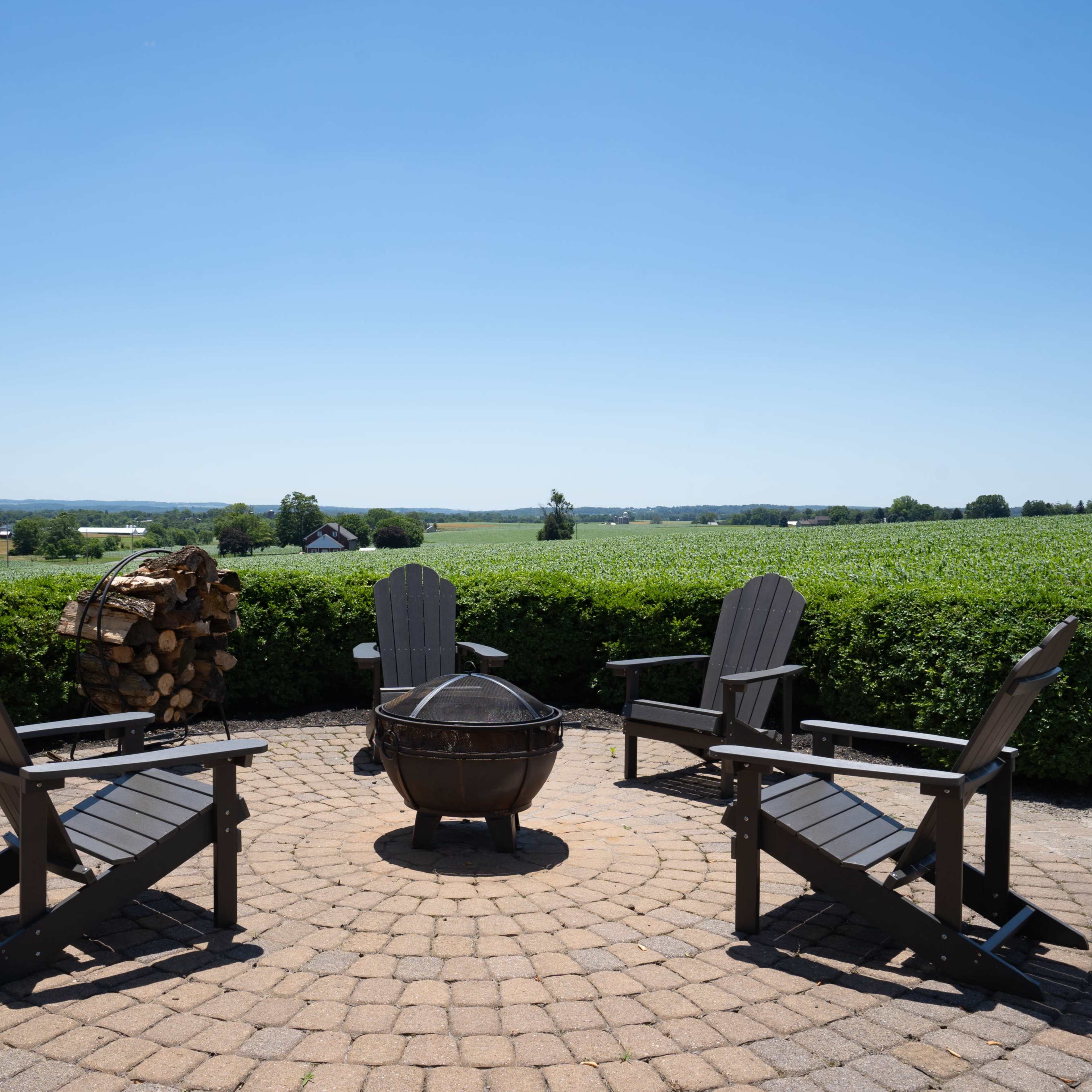 A circular patio with four Adirondack chairs surrounds a fire pit, set against a backdrop of farmland and a blue sky.