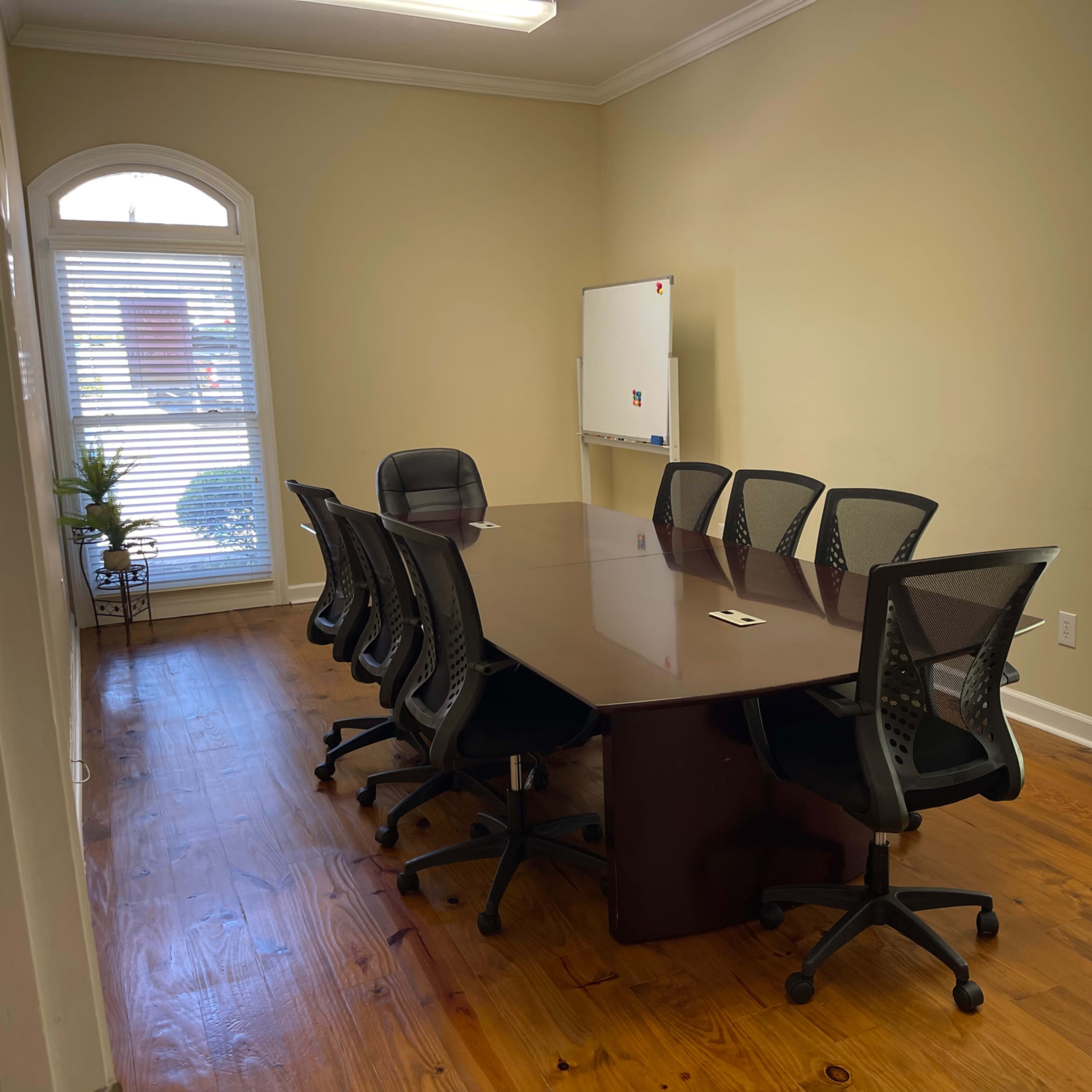 A conference room features a large wooden table surrounded by eight ergonomic chairs, with a whiteboard on the wall and windows letting in natural light.