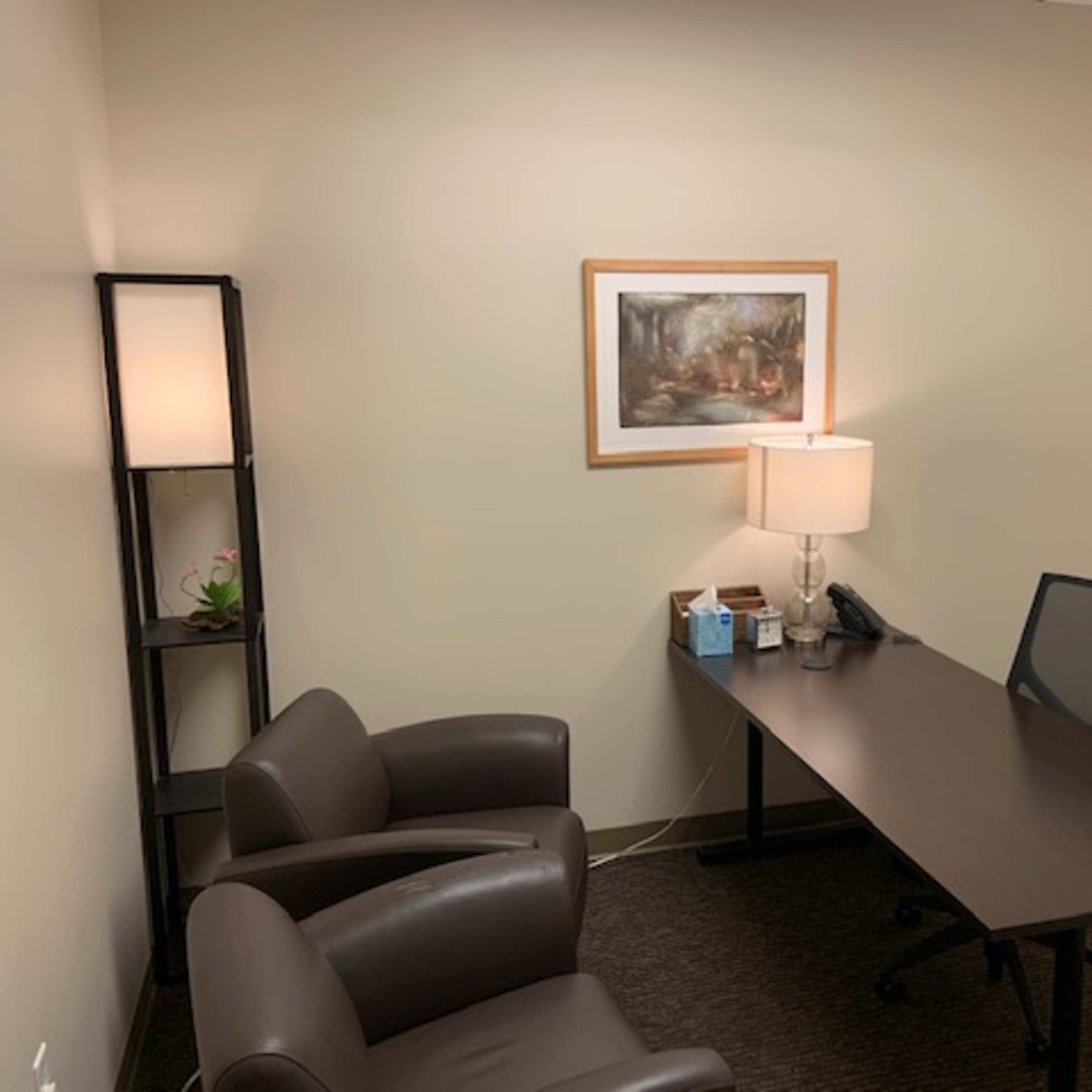 A small office with a desk, a chair, a lamp, and a framed artwork on the wall.