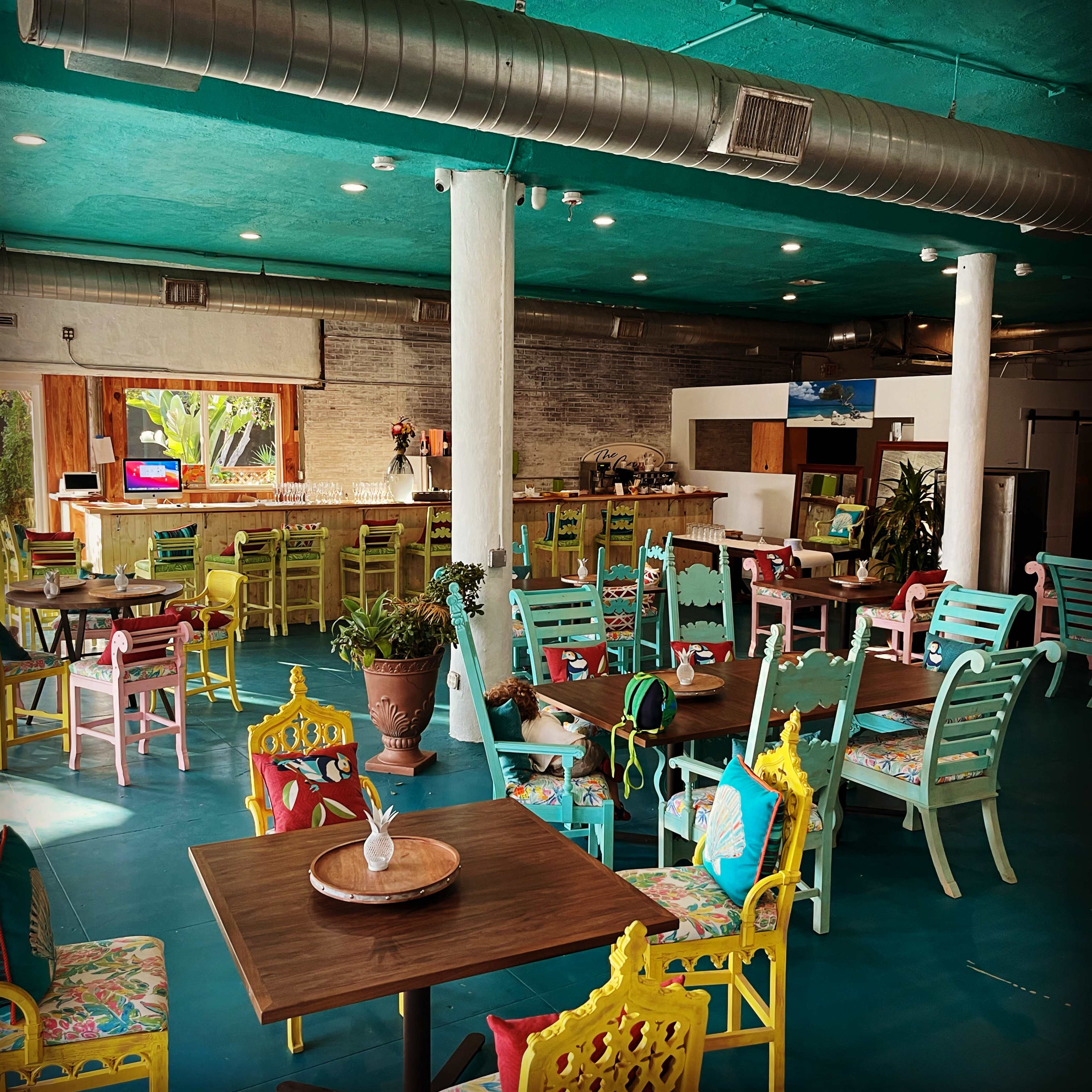 A brightly colored restaurant interior with wooden tables and chairs arranged around a spacious area, featuring a bar in the background and tropical decor.