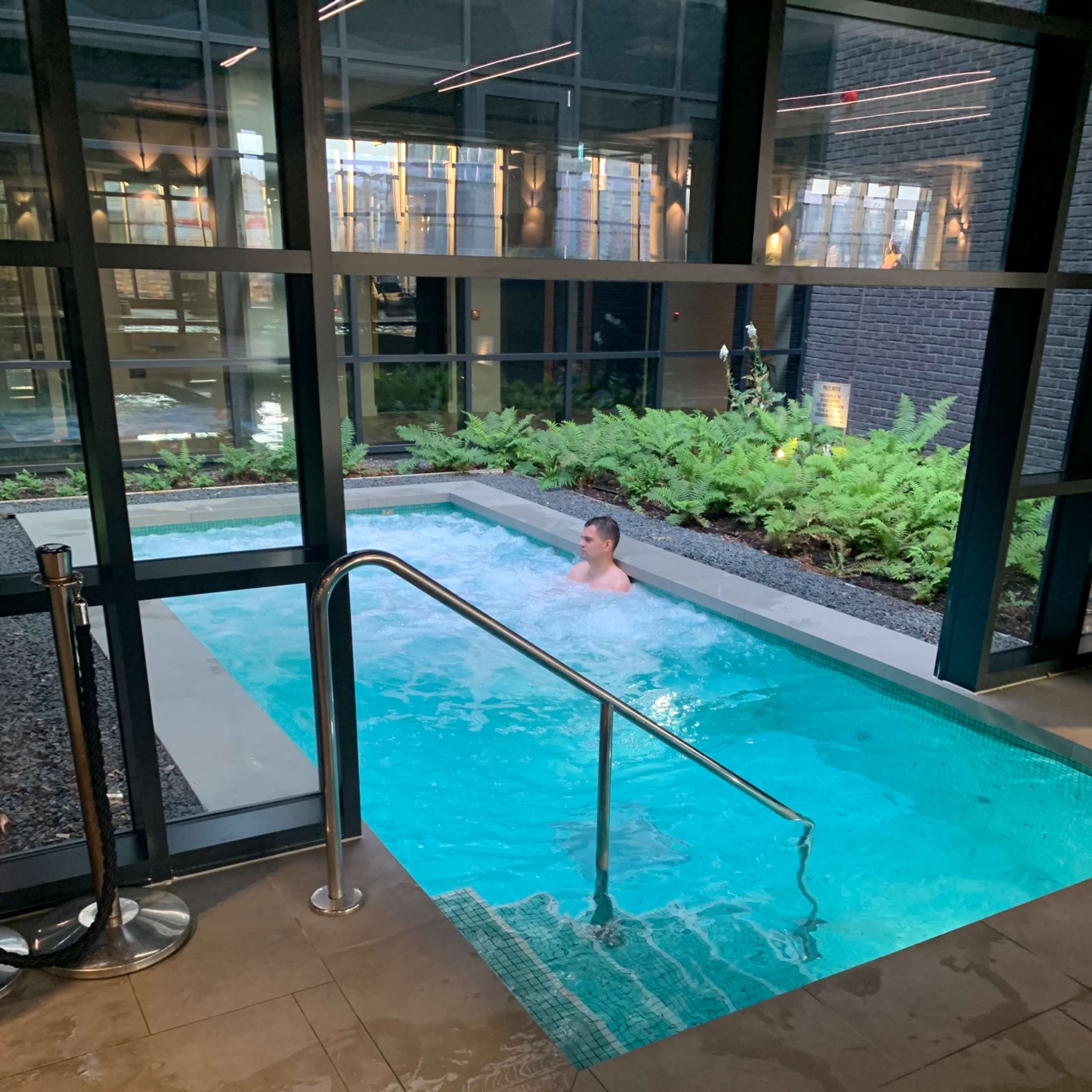 A person is swimming in a small indoor pool surrounded by large windows and greenery.