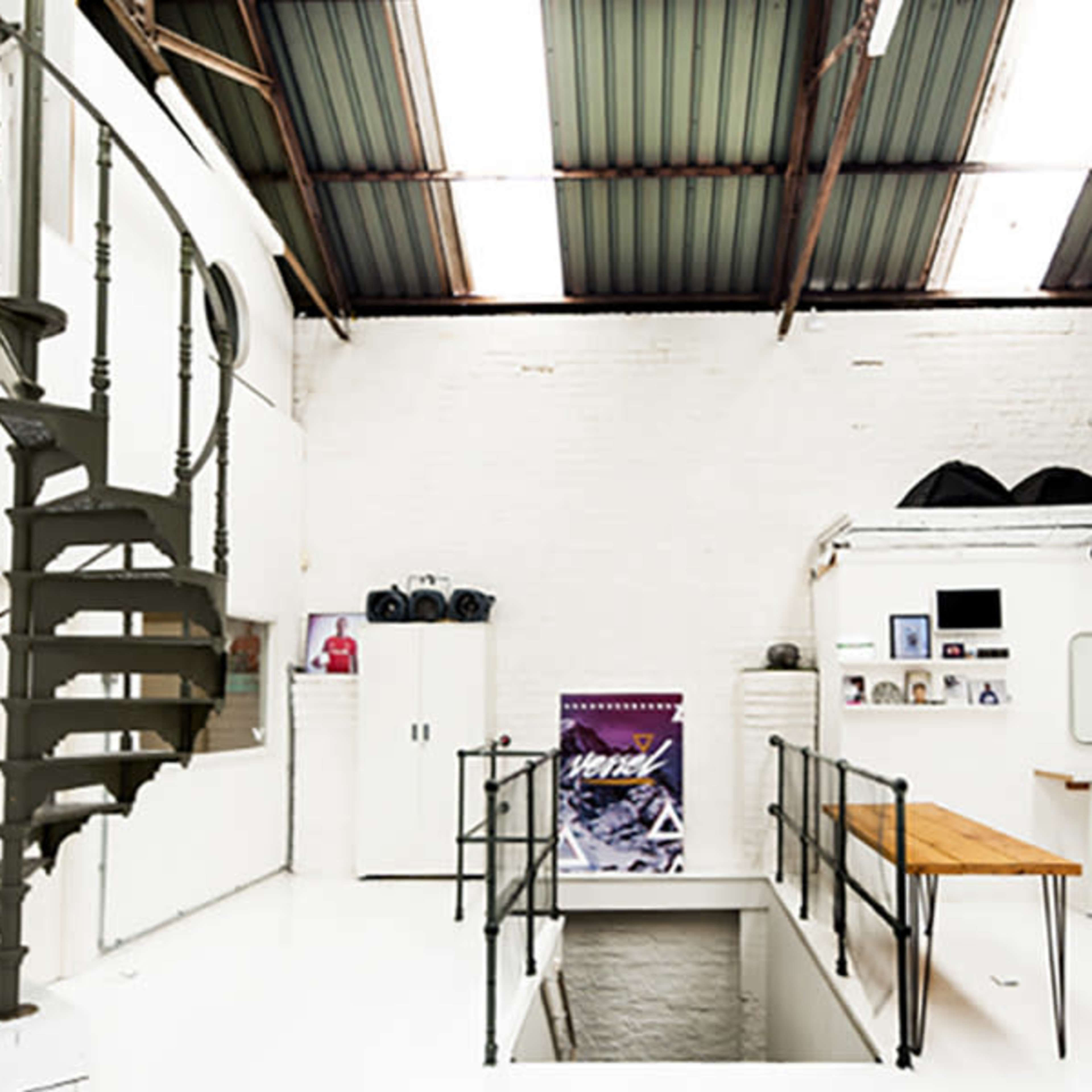 A modern, industrial-style studio with a spiral staircase, white walls, and various furniture pieces, including barstools and a long wooden table.