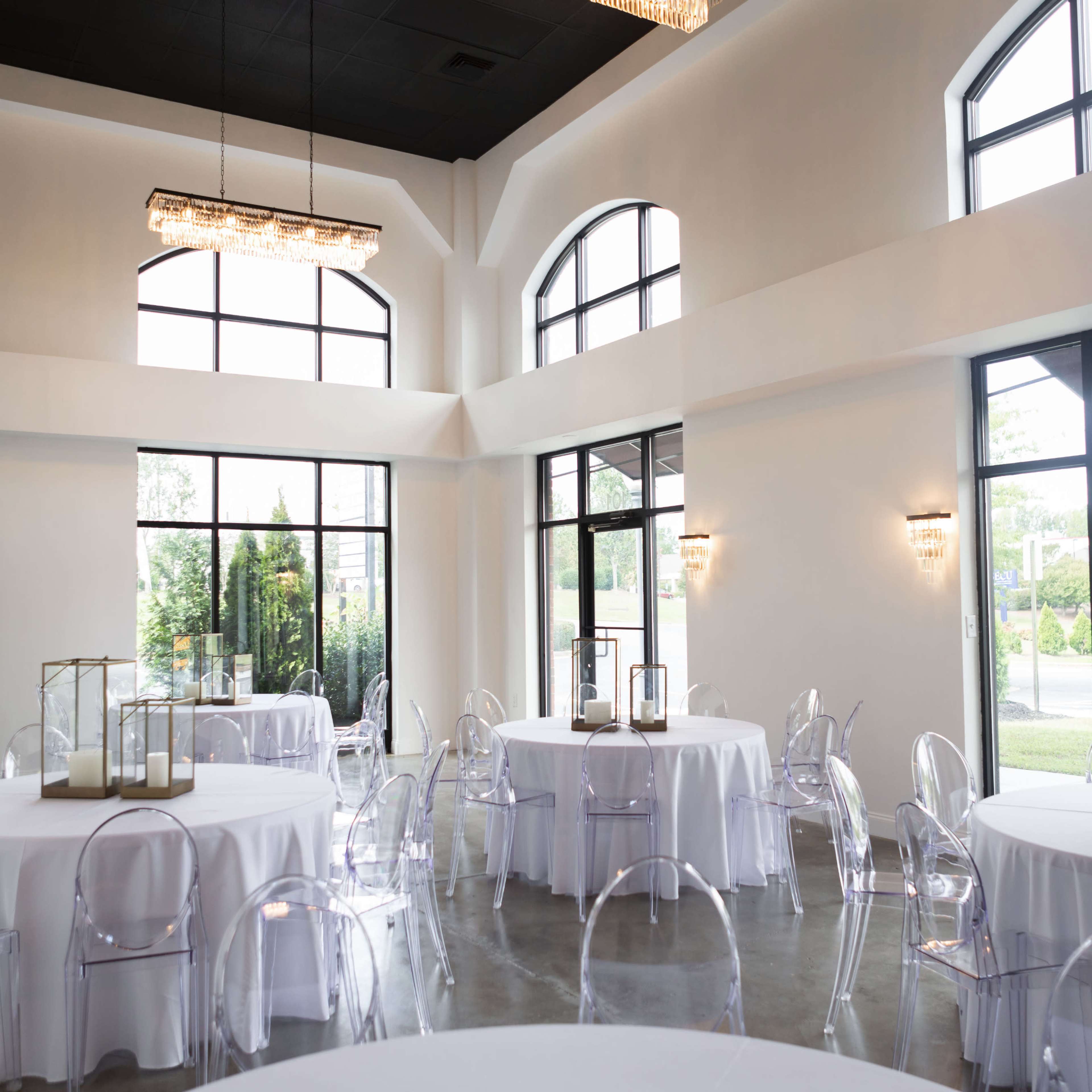 A bright event space with several round tables set, clear chairs, and large windows allowing natural light.