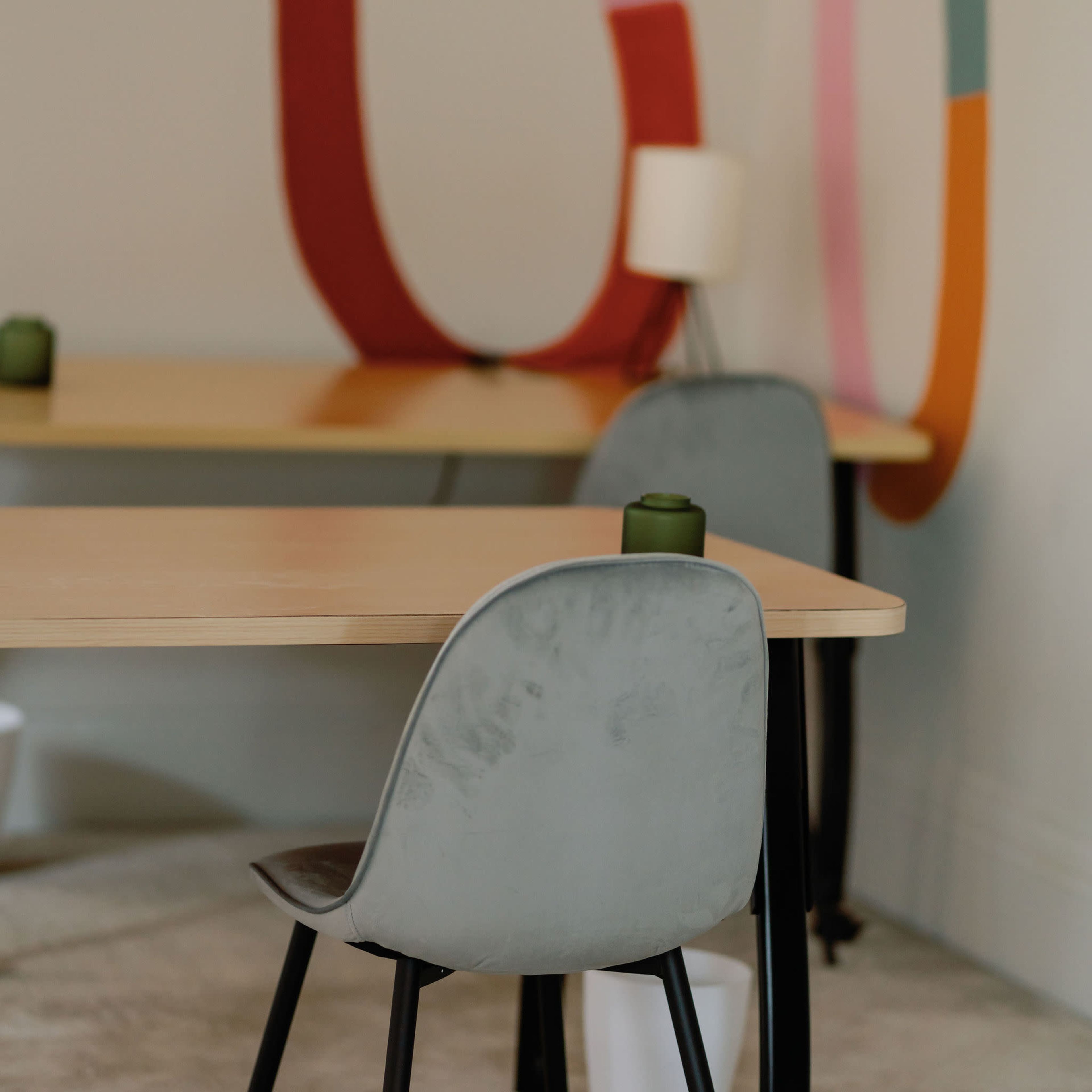 A minimalist interior with two wooden tables, each accompanied by a gray chair, and a colorful wall mural in the background.