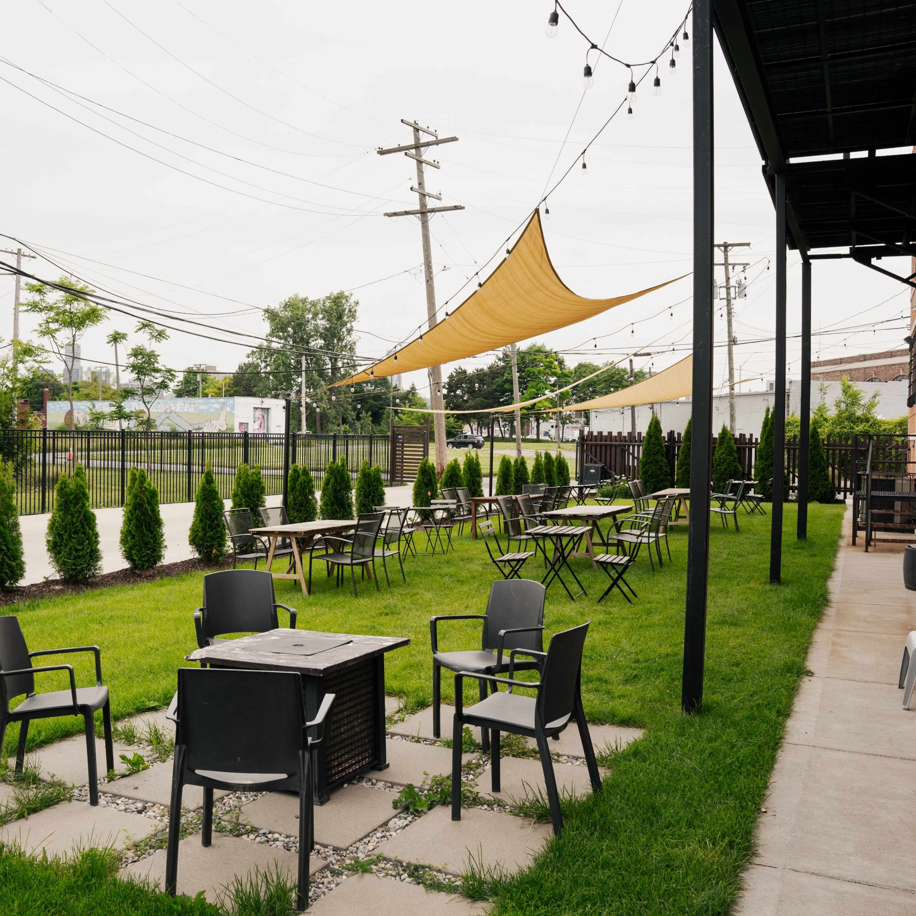 An outdoor seating area with metal chairs and tables surrounded by neatly arranged hedges and a large canopy providing shade.