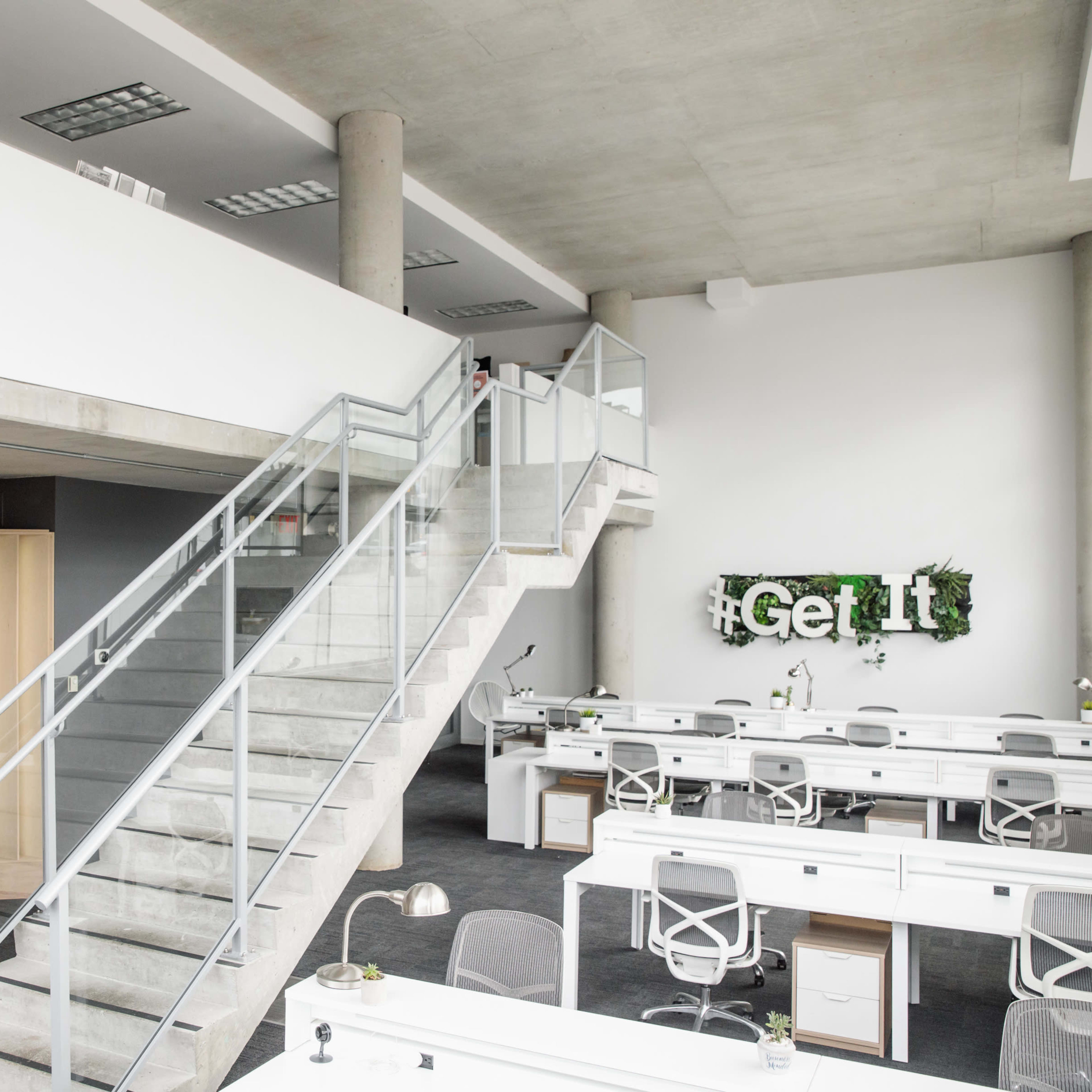 A modern office space with a staircase, a "TALK" sign on a wooden wall, and several workstations equipped with desks and plants.