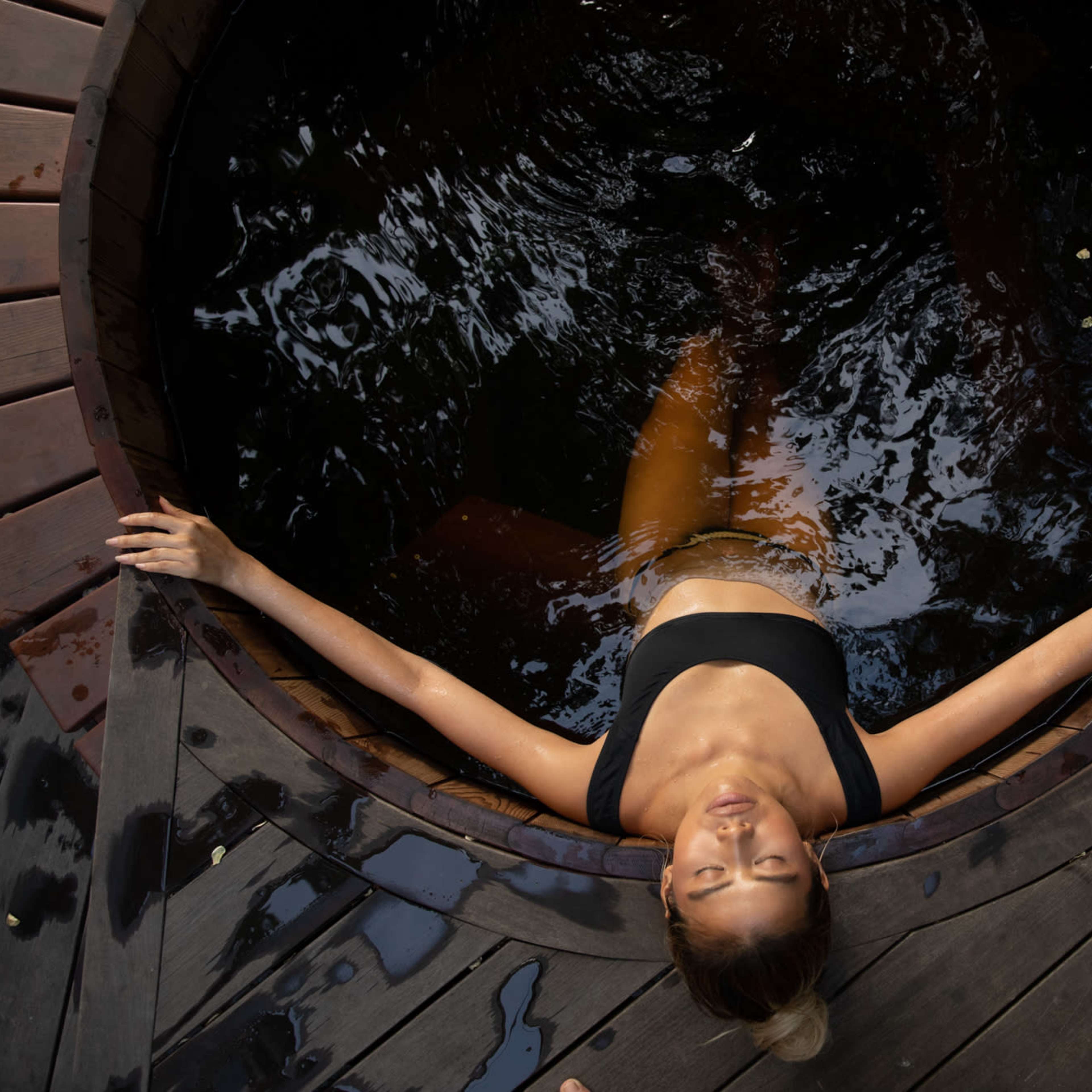A woman relaxes in a wooden hot tub surrounded by dark water and a wooden deck.