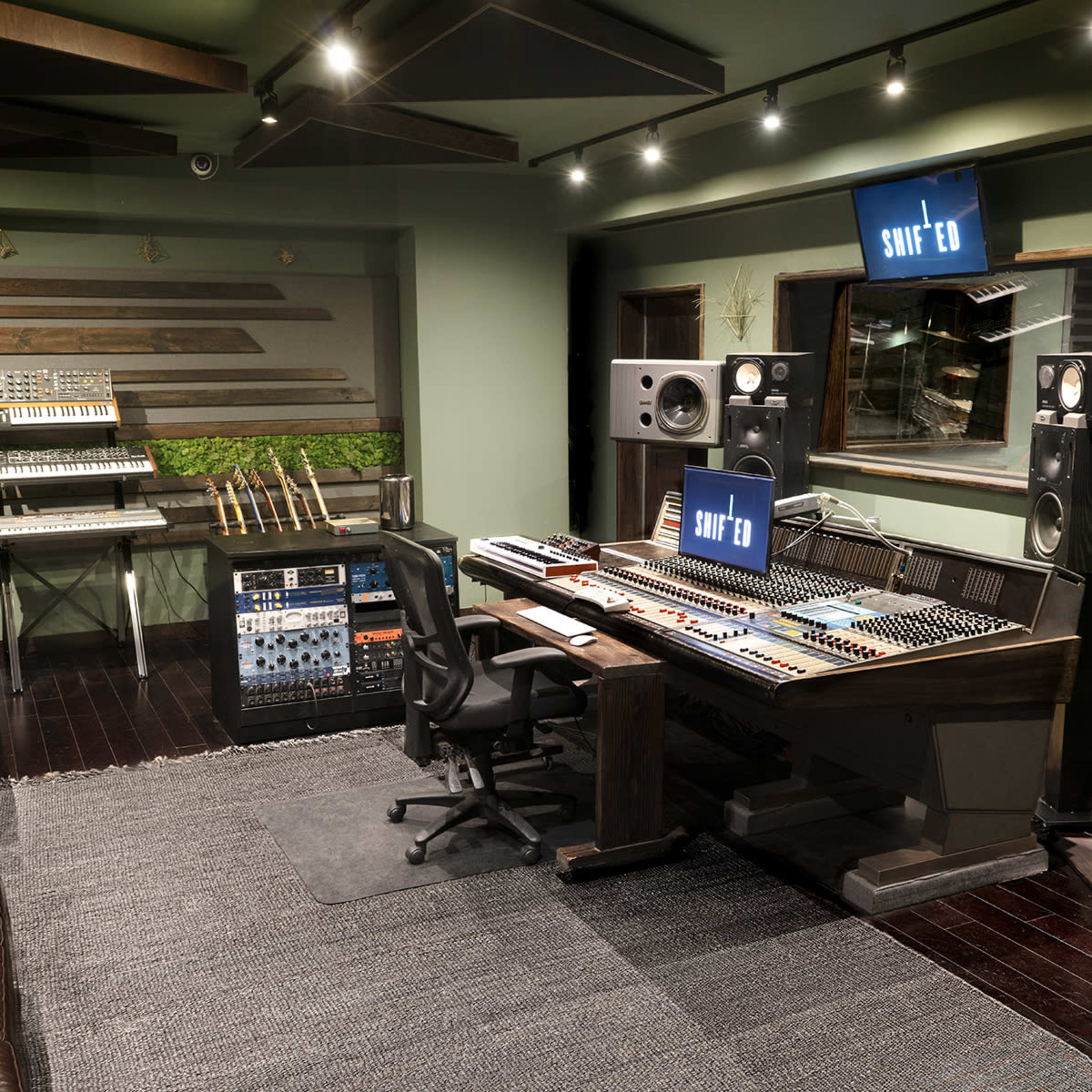 A recording studio featuring a large mixing console, audio equipment racks, and various musical instruments, with green walls and ambient lighting.