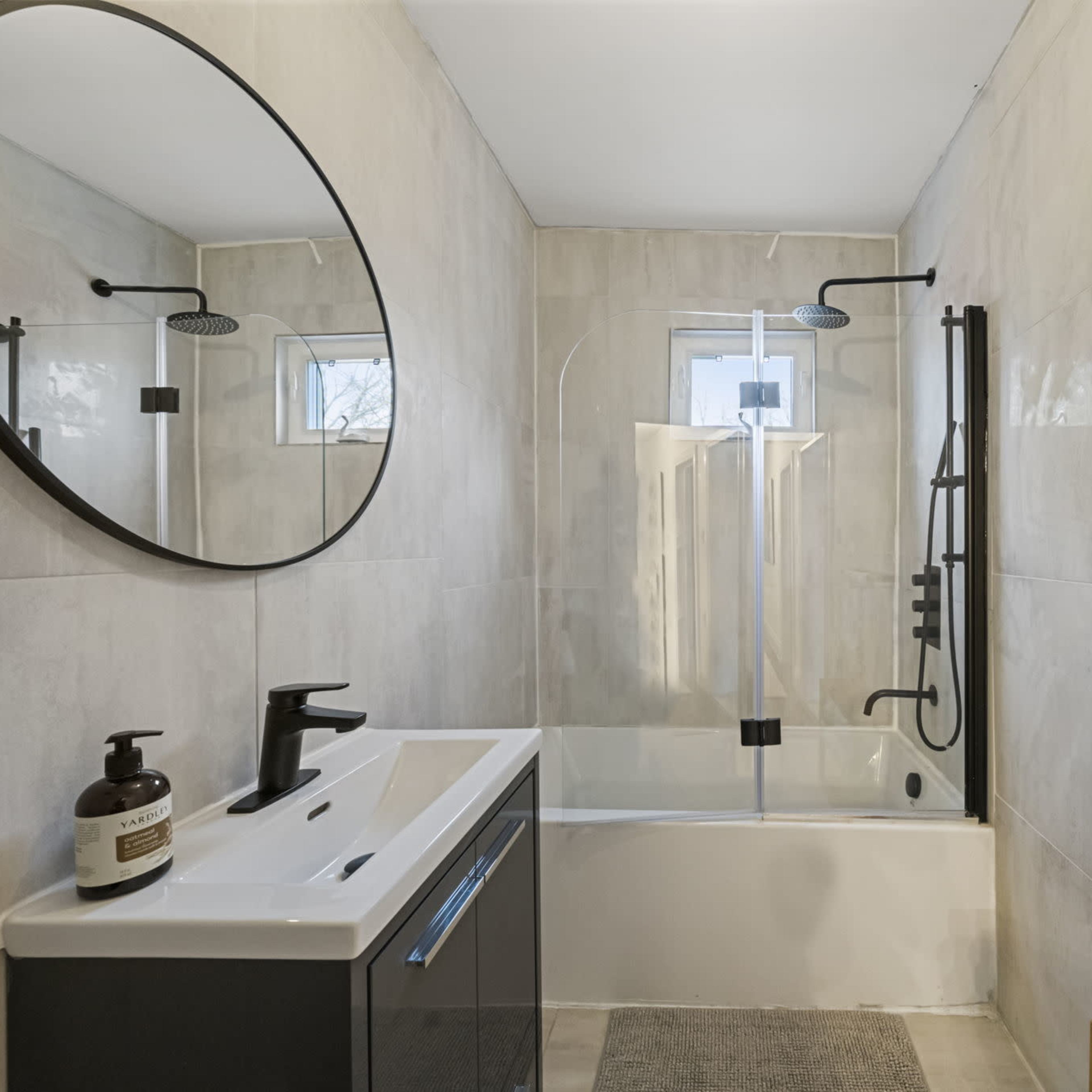 A modern bathroom featuring a bathtub with a glass shower panel, a circular mirror above a dark vanity, and gray tiled walls.