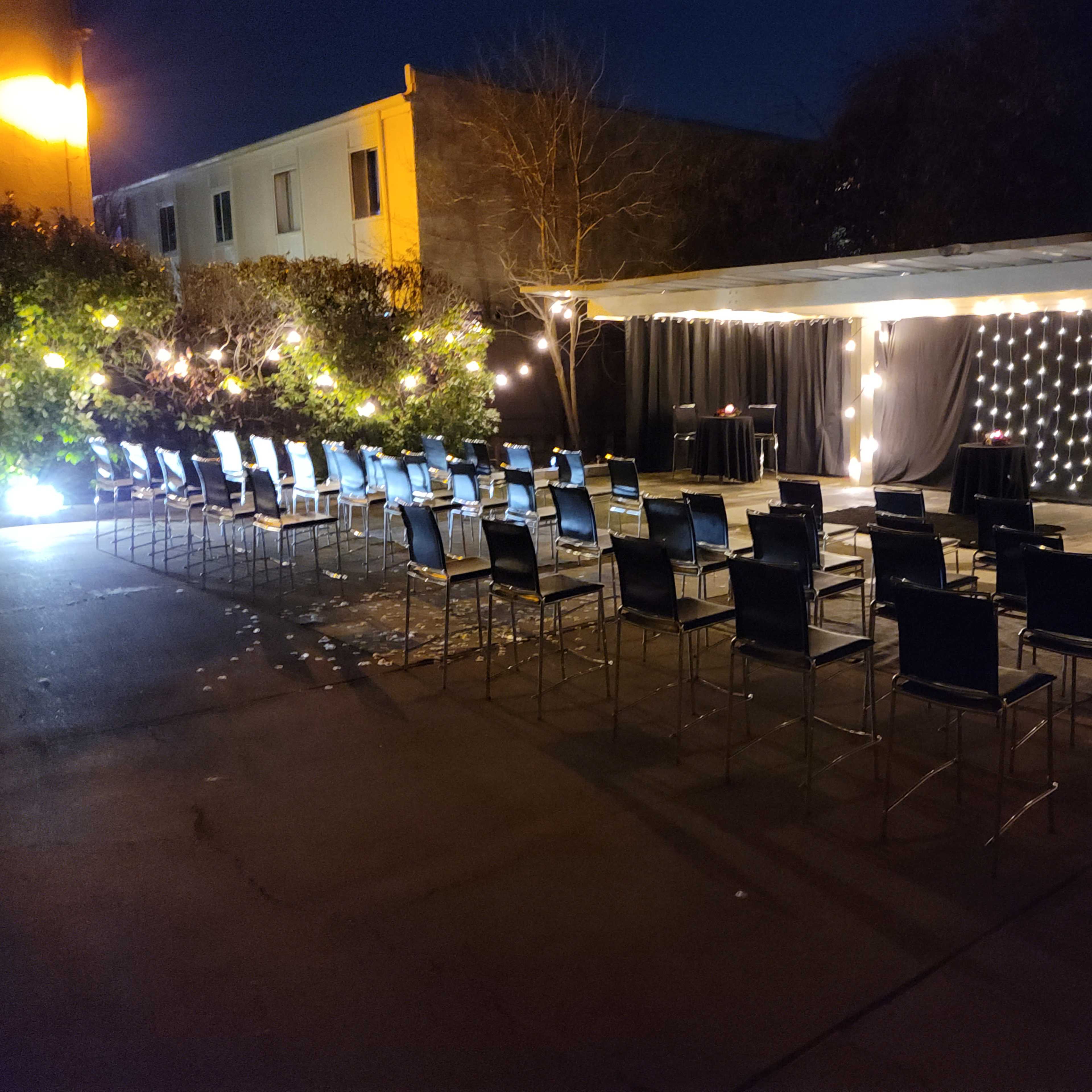 A dimly lit outdoor venue set up with rows of black chairs facing a decorated backdrop and string lights.
