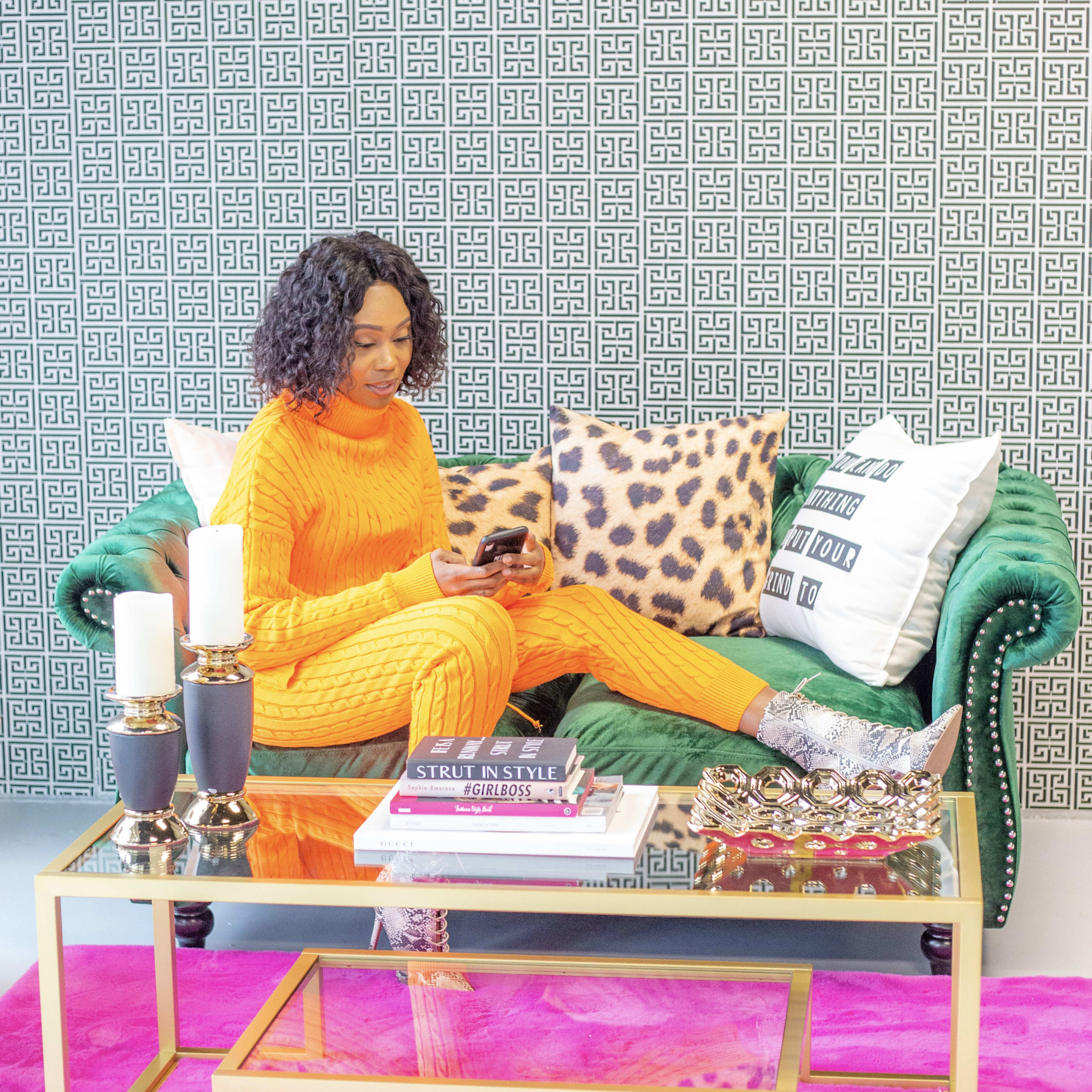 A woman in an orange outfit sits on a green couch, using her phone, surrounded by decorative pillows and a glass coffee table.
