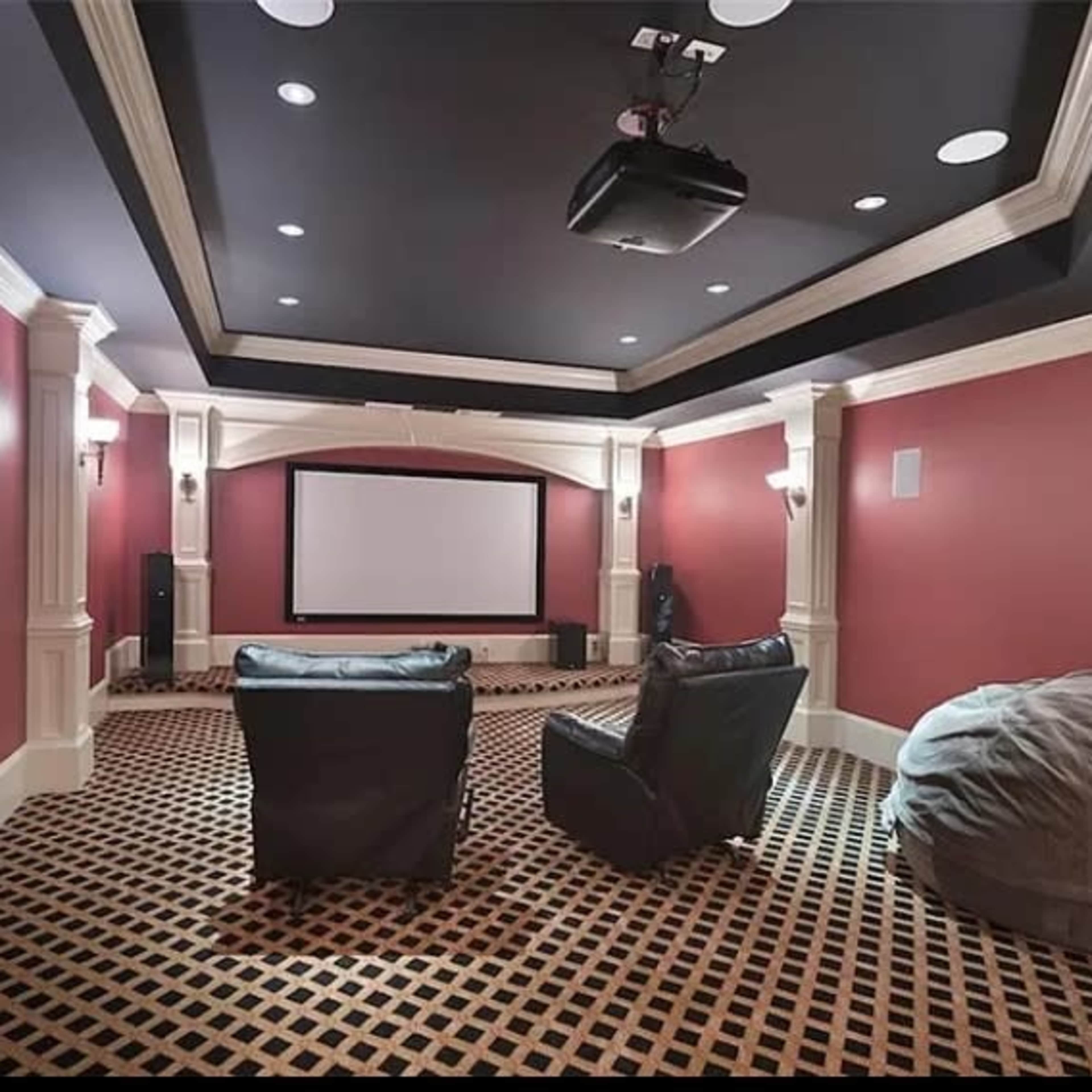 A home theater room with a large screen at the front, two leather chairs, a bean bag, and red walls adorned with decorative moldings.