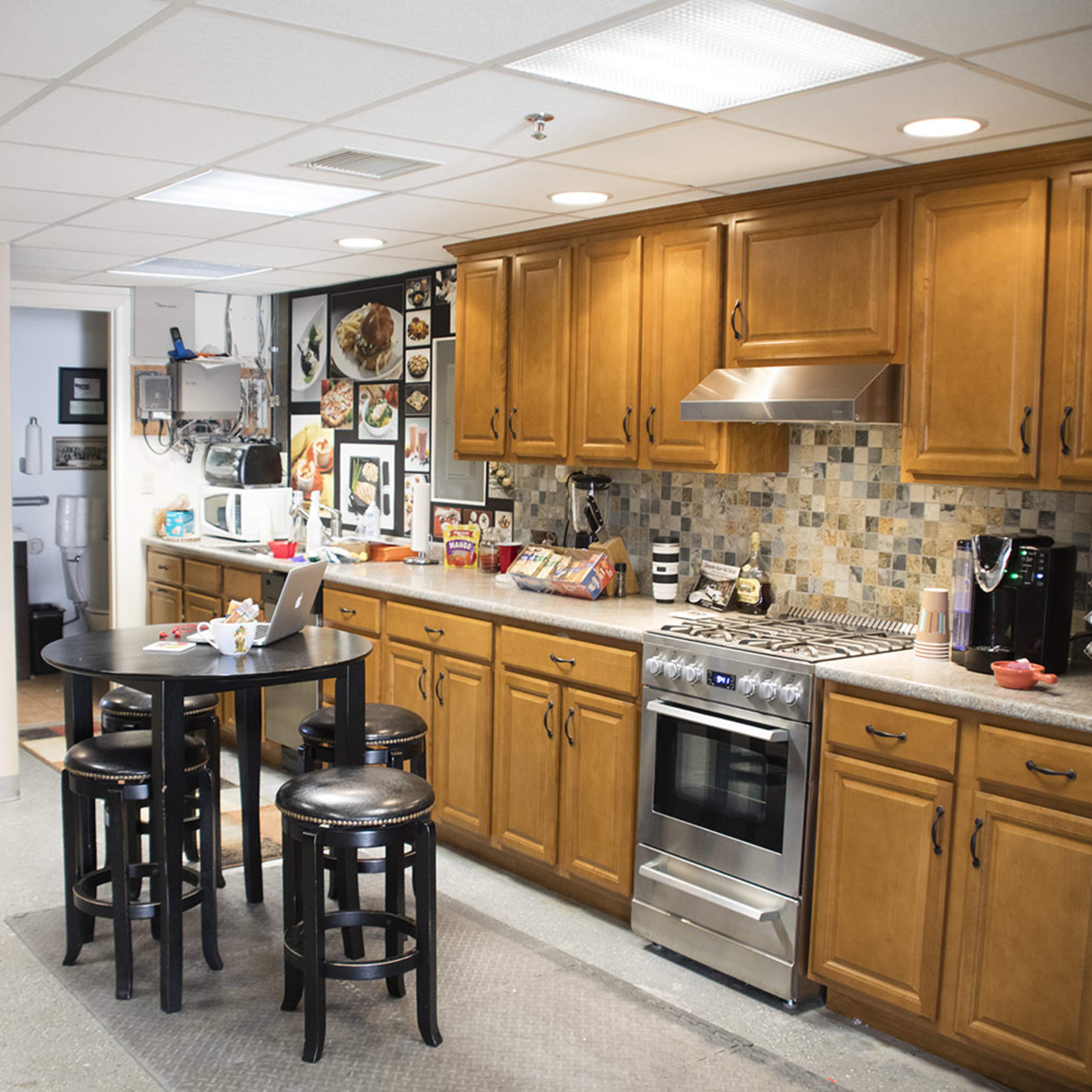 A modern kitchen with wooden cabinets, a countertop, a stove, and a table with black stools, adjacent to a hallway leading to another room.