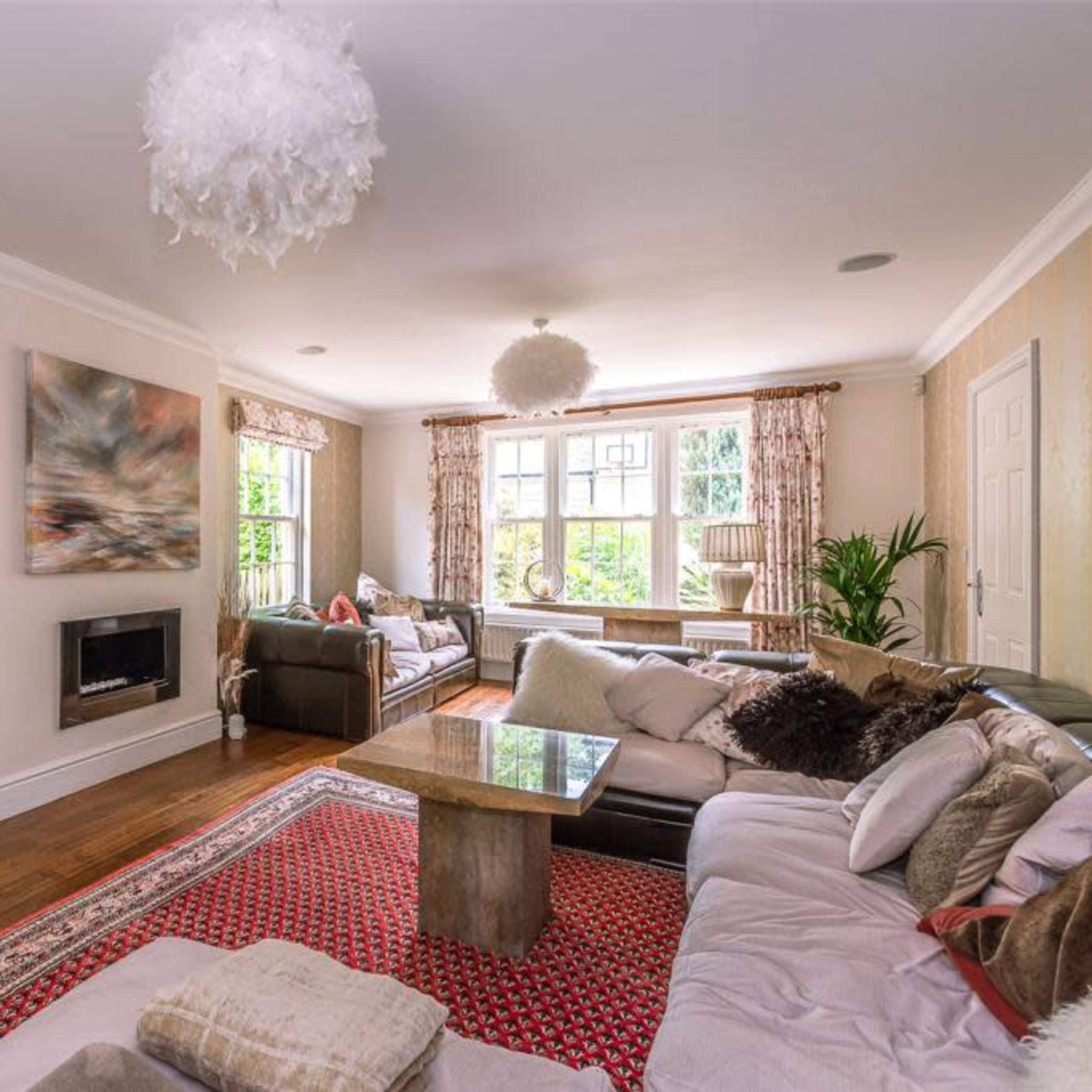 The living room features a large area rug, multiple couches, a mounted television, and a fireplace, with natural light streaming in through large windows.