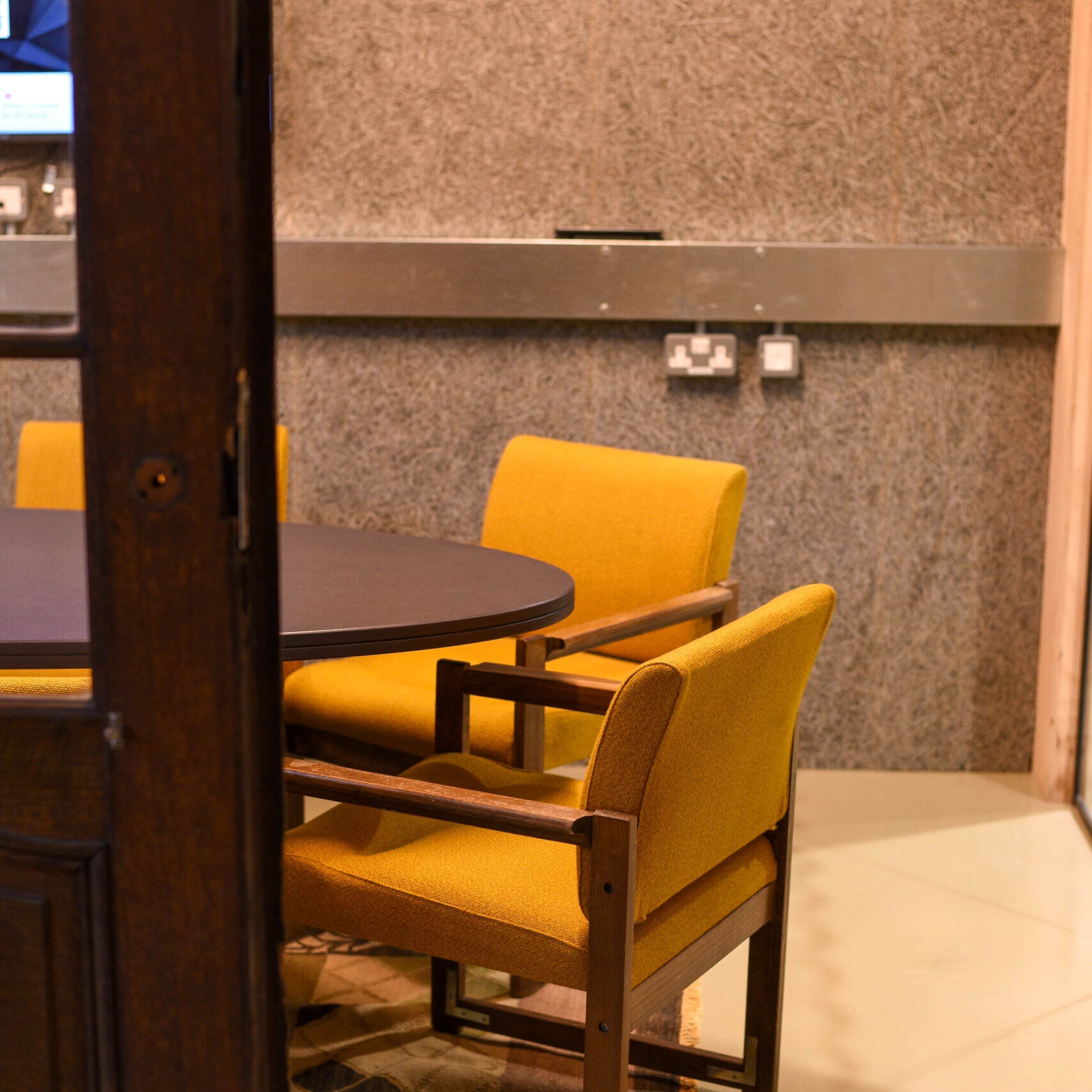 A wooden door partially opens to reveal a round table with yellow upholstered chairs on a patterned rug in a room with textured gray walls.