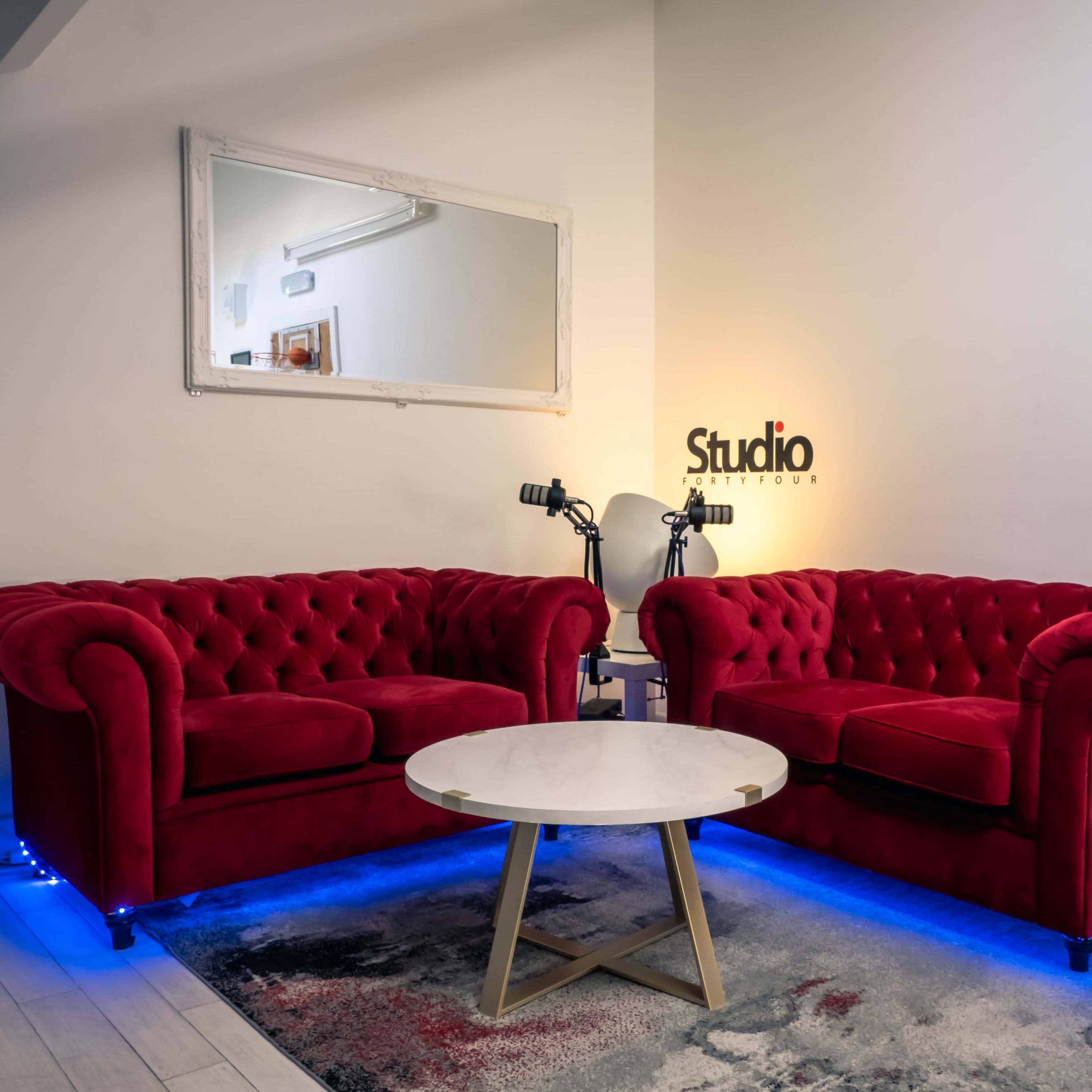 A pair of red tufted sofas are positioned opposite a small round table in a well-lit studio space with a mirror on the wall.