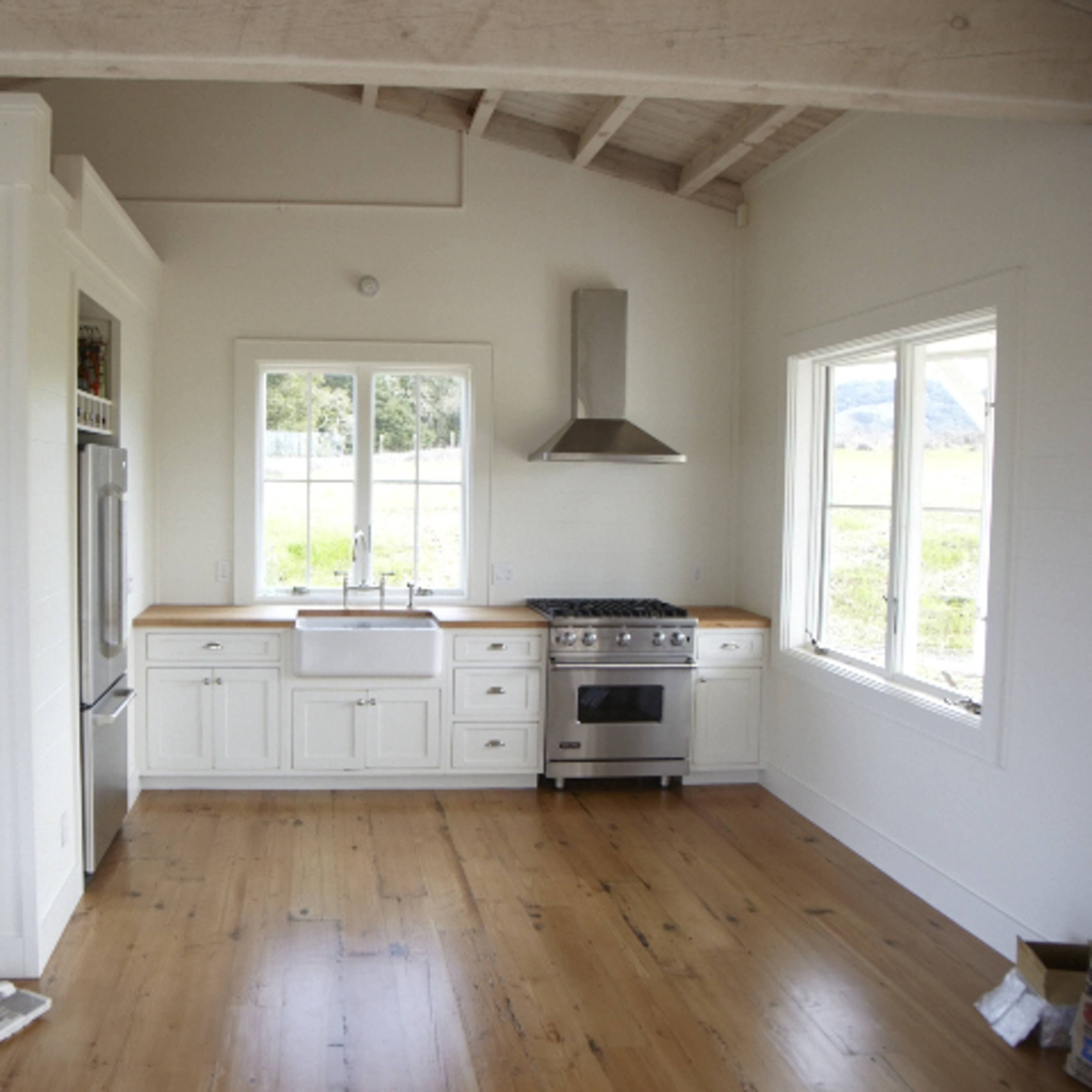 A modern kitchen with white cabinetry, a stainless steel stove, a farmhouse sink, and large windows letting in natural light.