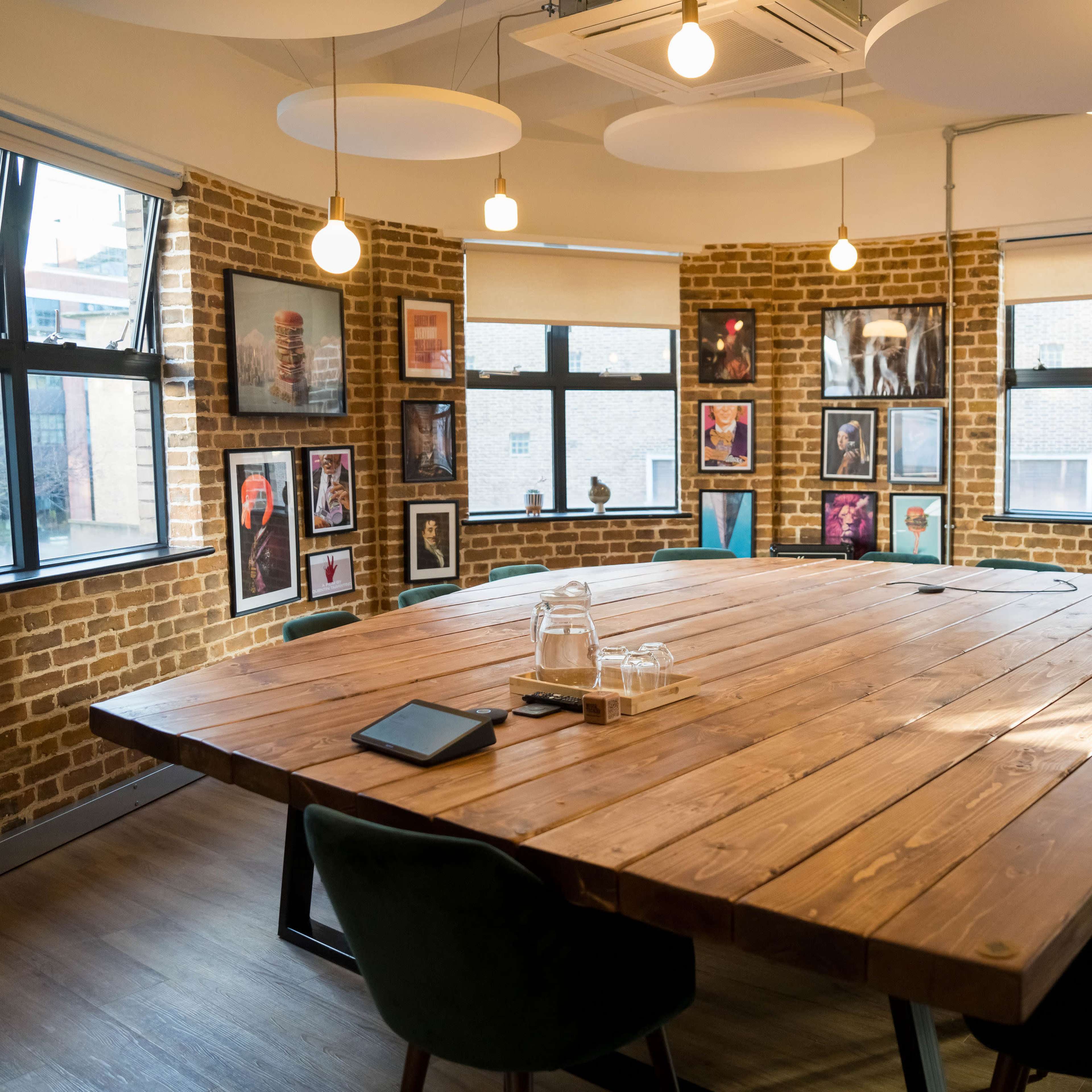 A large wooden conference table surrounded by green chairs is set in a room with brick walls and framed artwork on display.