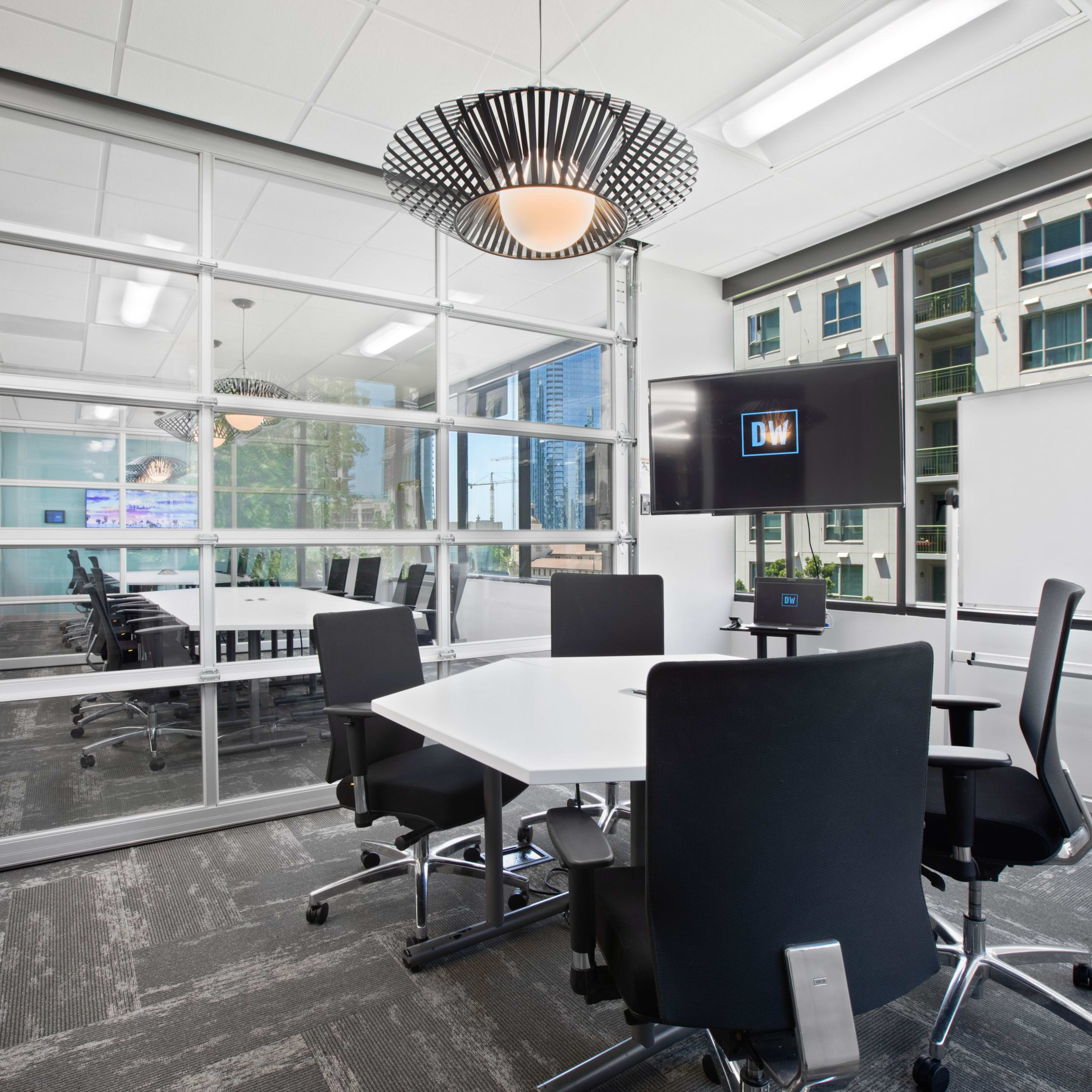 A modern conference room with a glass wall, a whiteboard, a television screen, and several black office chairs around a white table.