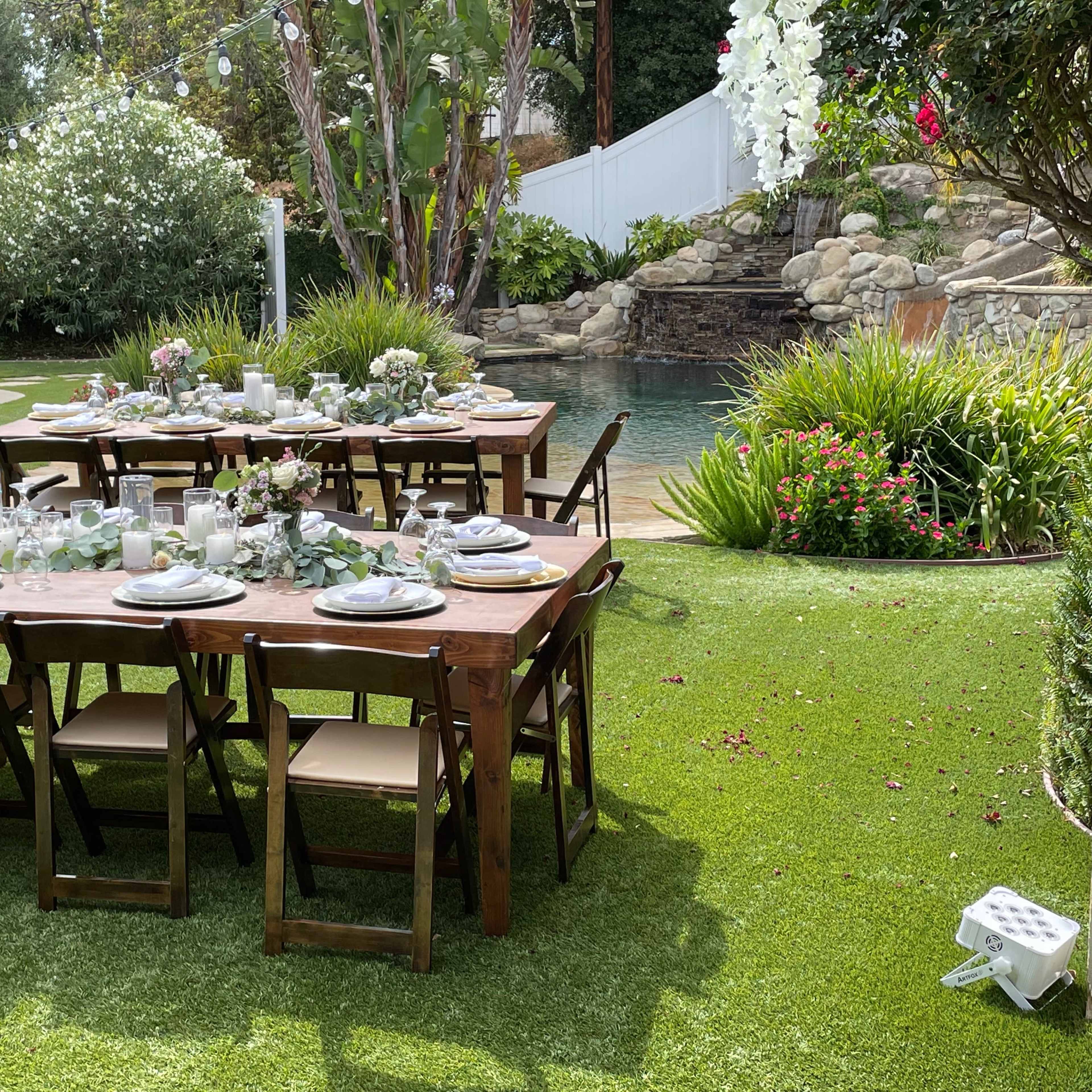 A long wooden dining table is set with plates and centerpieces in a lush garden near a pool.