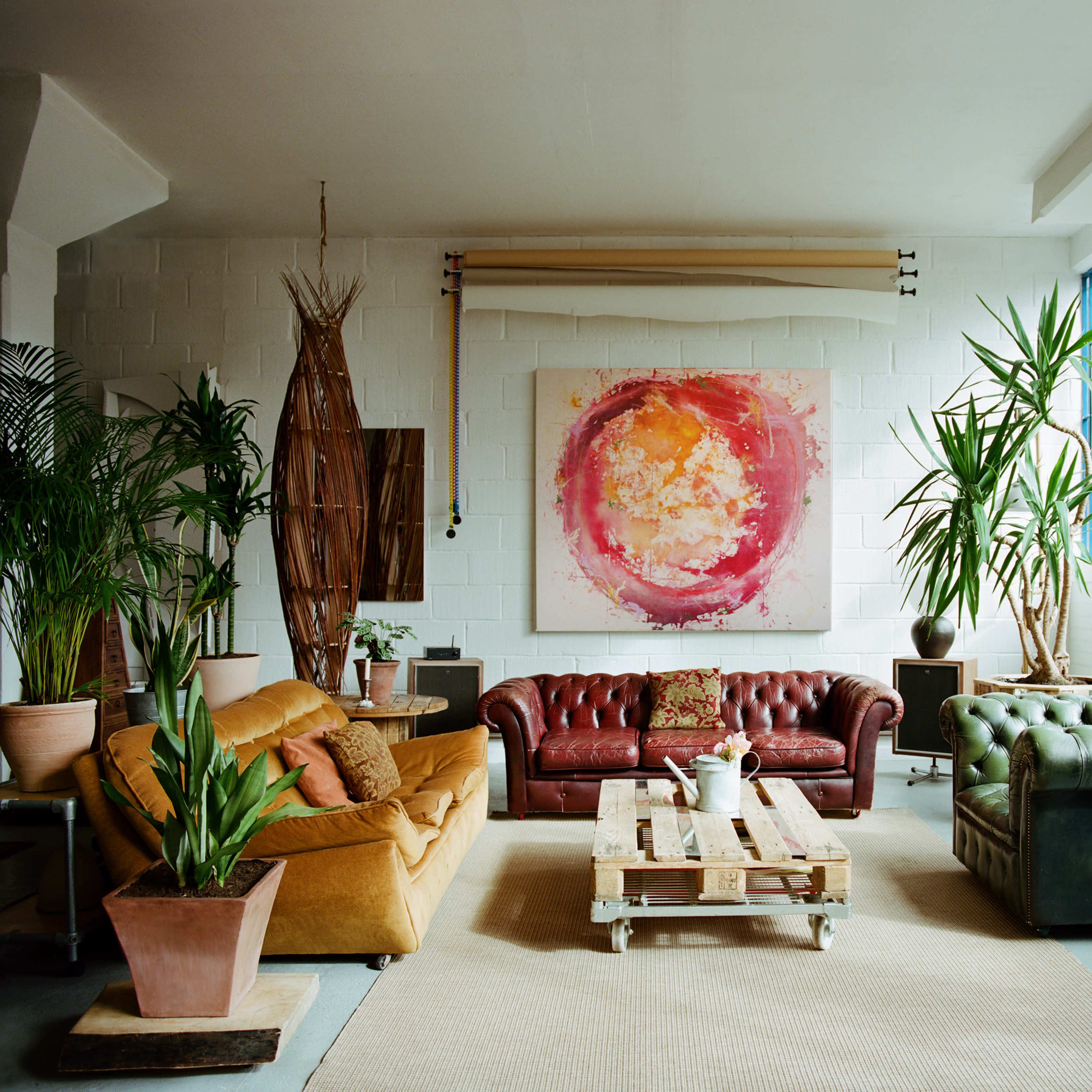 A spacious living room with a mix of leather sofas, a wooden coffee table made from pallets, and several potted plants beside large windows.