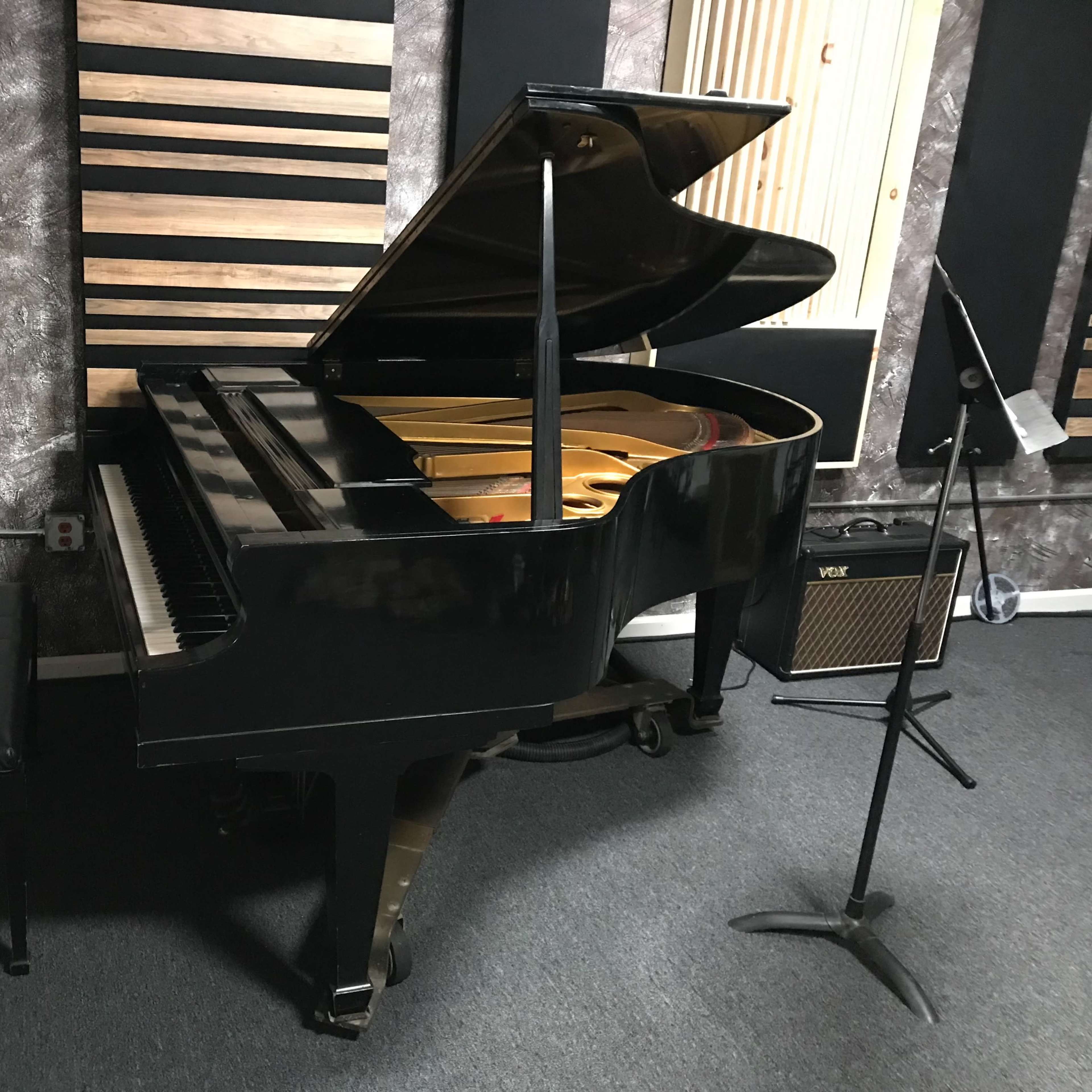 A black grand piano is positioned in a music studio with soundproof panels and a microphone stand nearby.