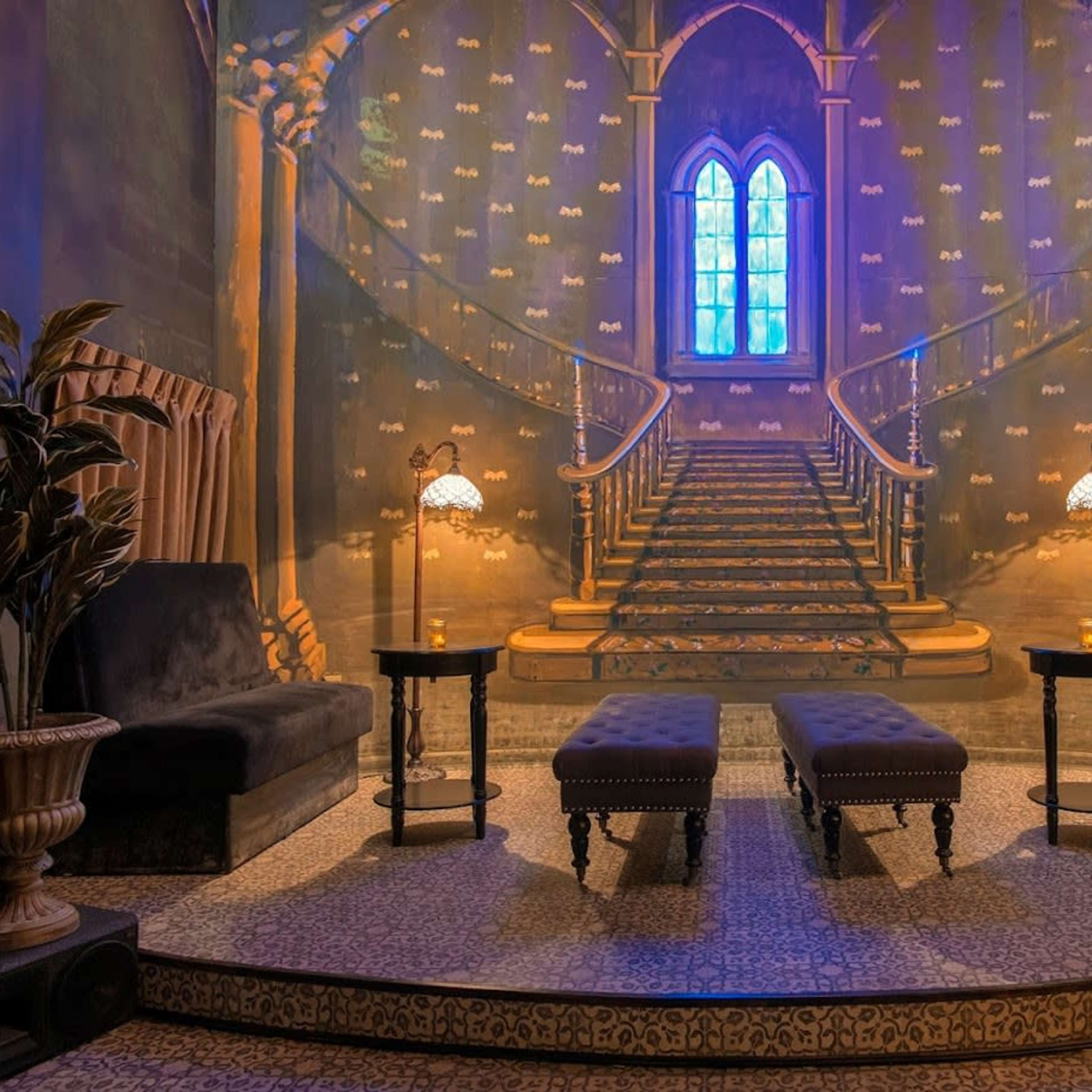 A dimly lit interior with plush seating arranged in front of a large mural depicting a grand staircase and a stained glass window.