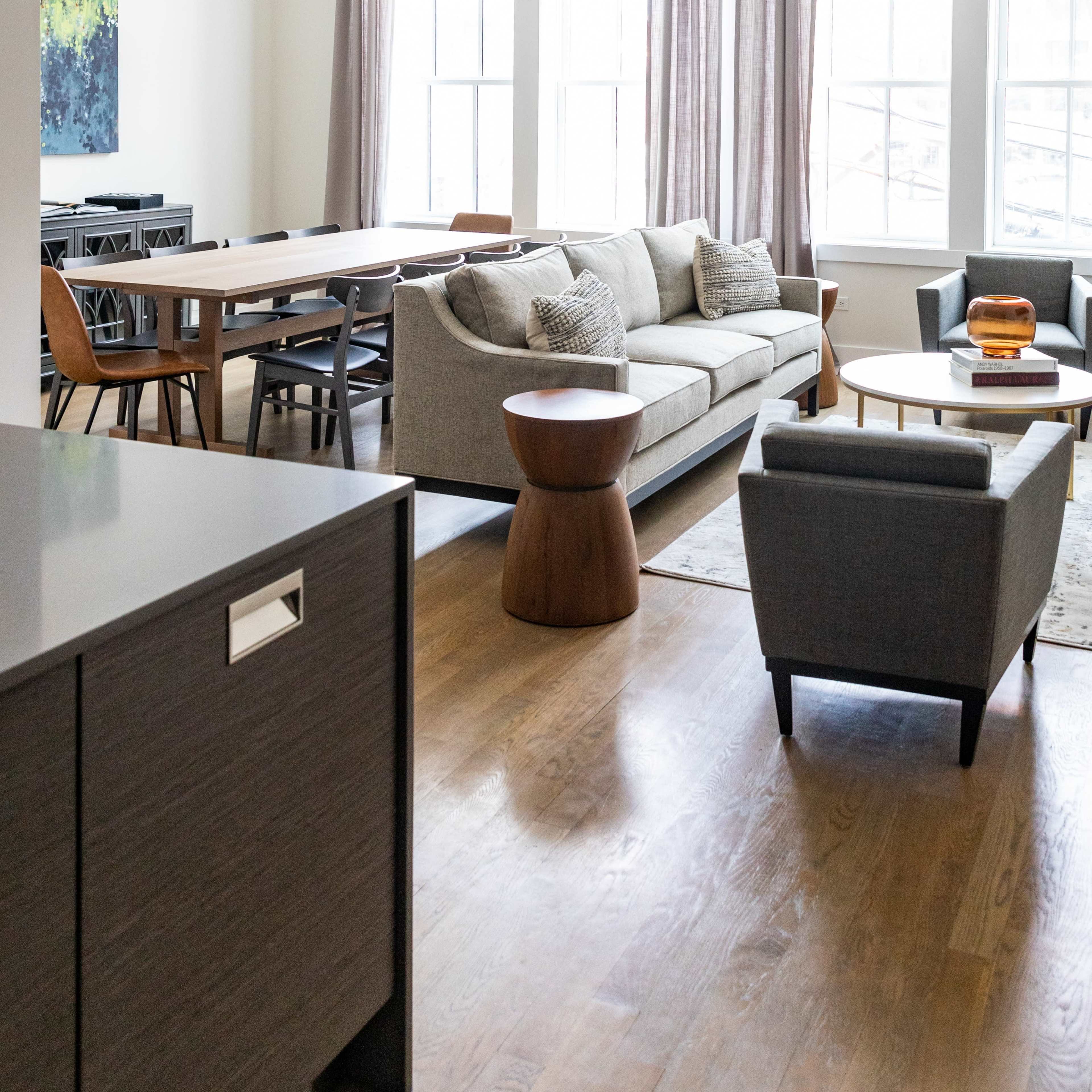 A modern living area featuring a sofa, armchairs, a coffee table, and a dining table with chairs, all arranged around large windows with sheer curtains.
