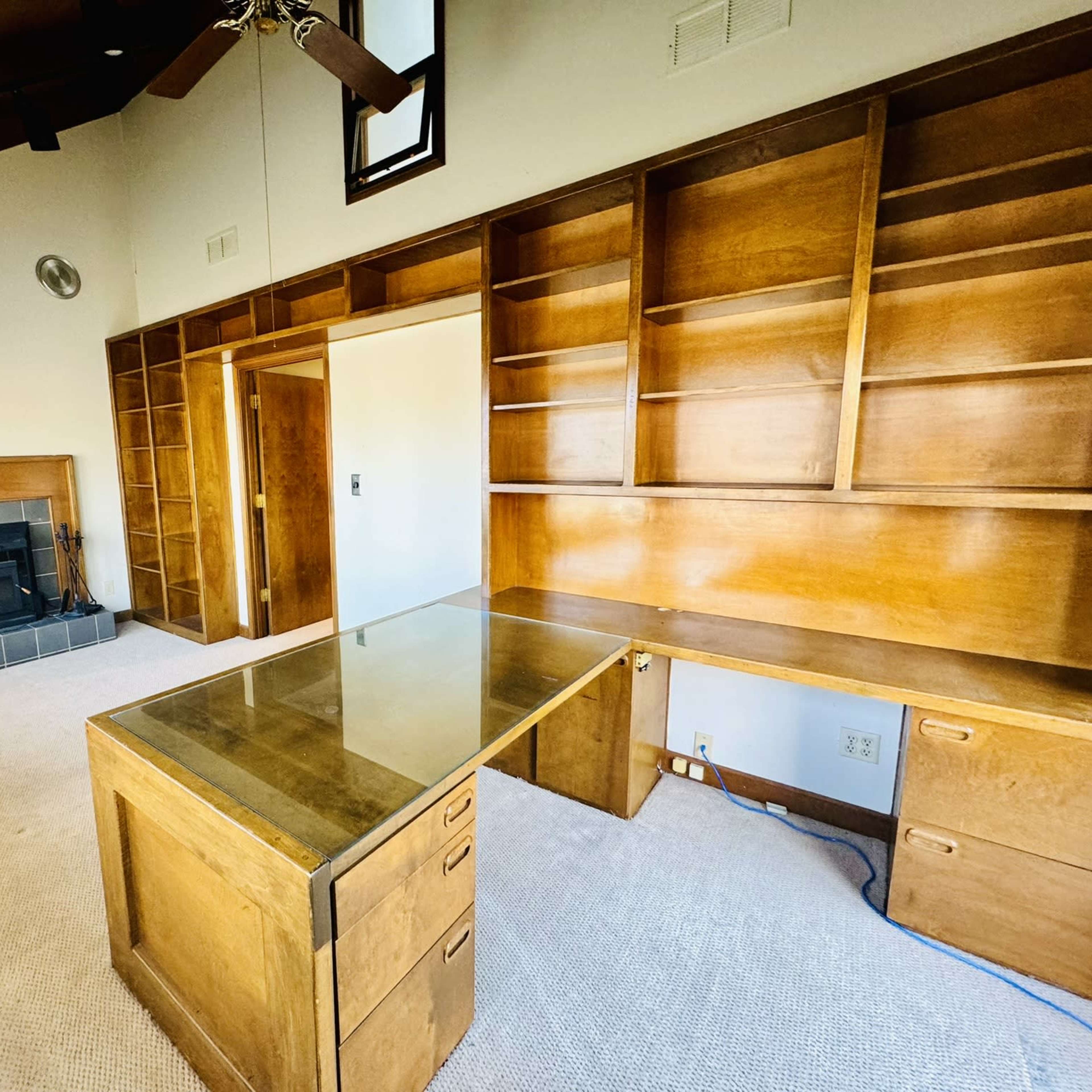 A room with a wooden desk and shelving unit against a wall, featuring a fireplace and a high ceiling.