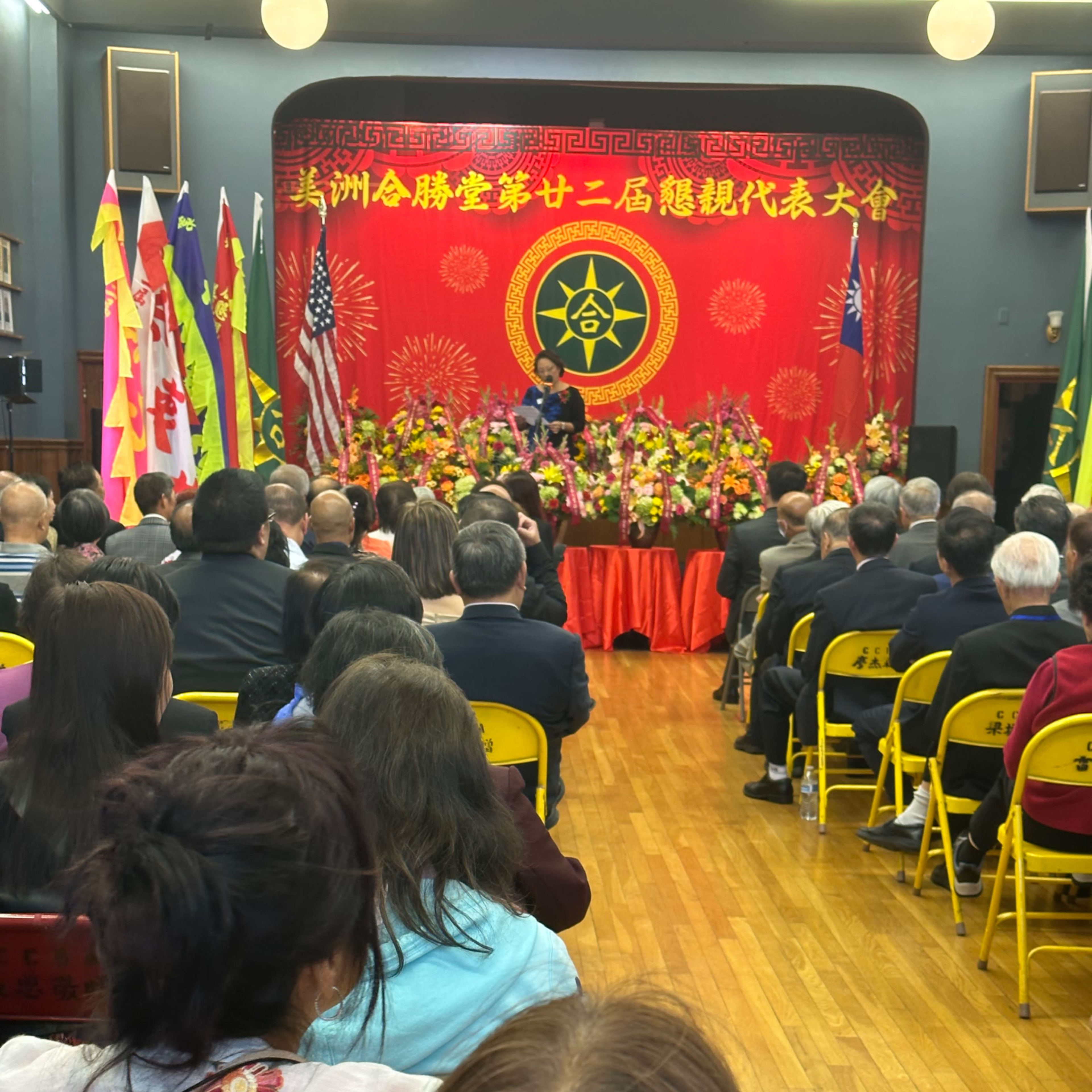 A speaker addresses an audience at a formal event decorated with flags and flowers in a venue featuring a red backdrop.