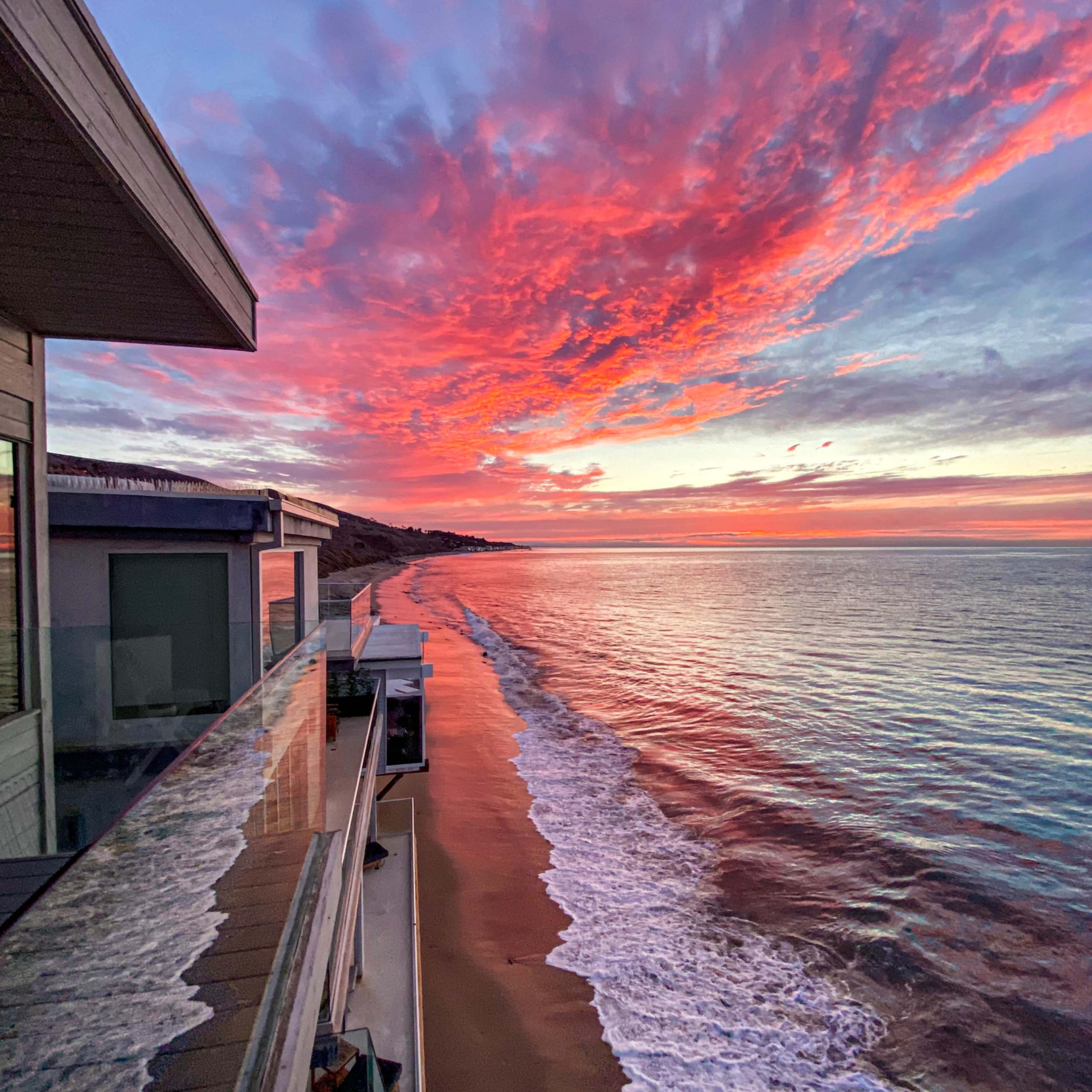 A vibrant sunset casts colorful reflections over a calm ocean along a sandy beach, with a modern building on the left.
