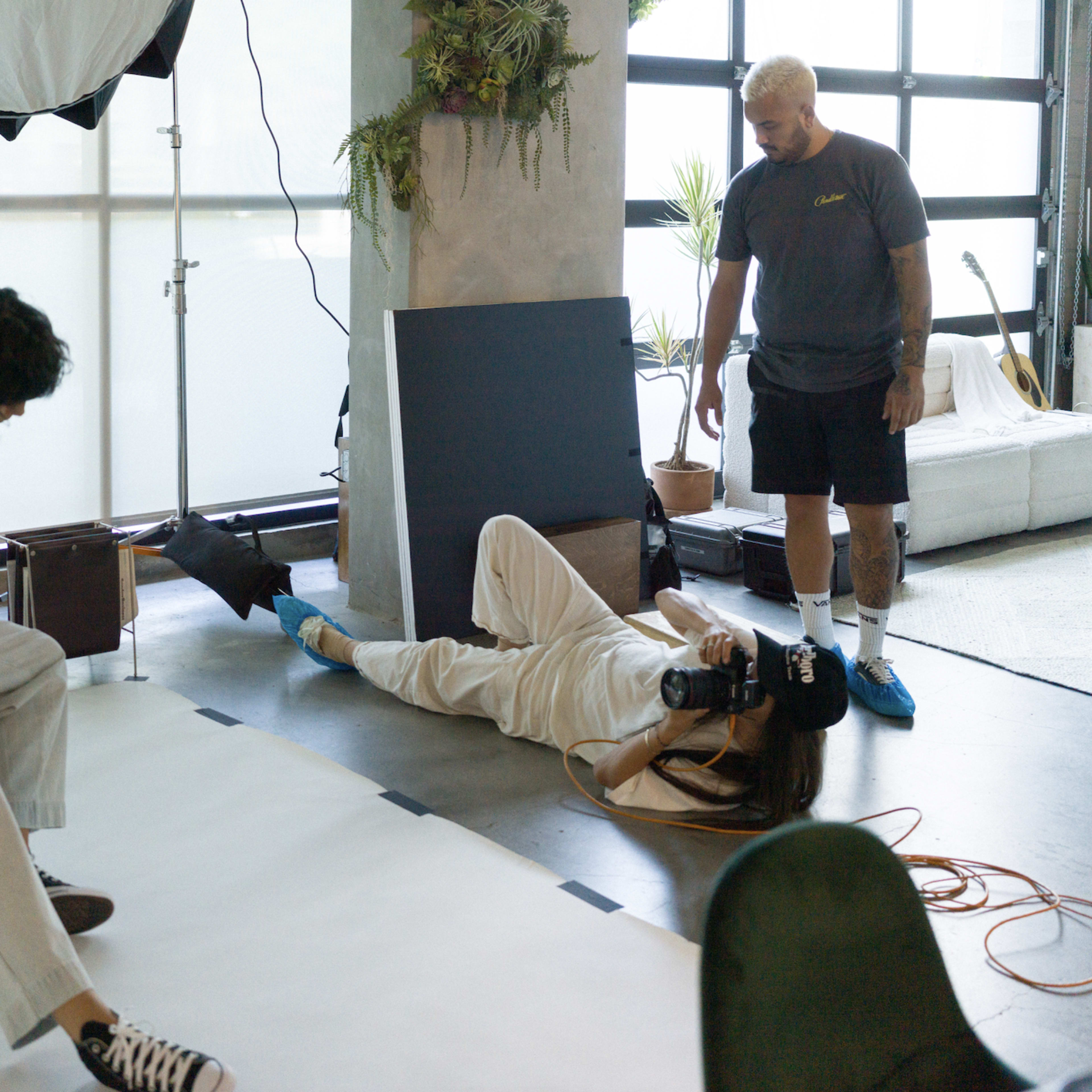 A photography studio where a model sits on a wooden stool while another model lies on the floor, posing for a photographer.
