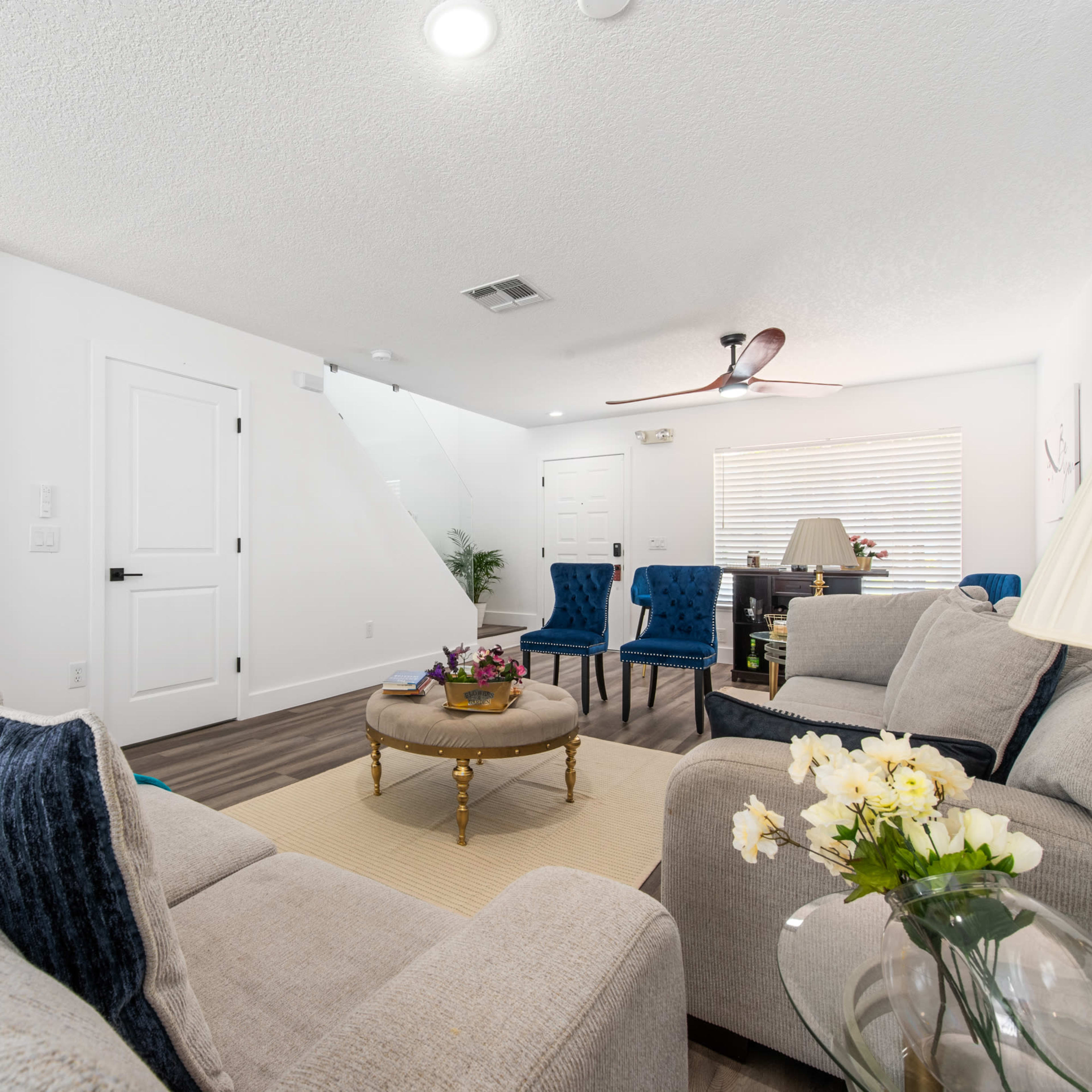 A modern living room with neutral-colored sofas, a round coffee table, and blue upholstered chairs, featuring a ceiling fan and natural light from a window.
