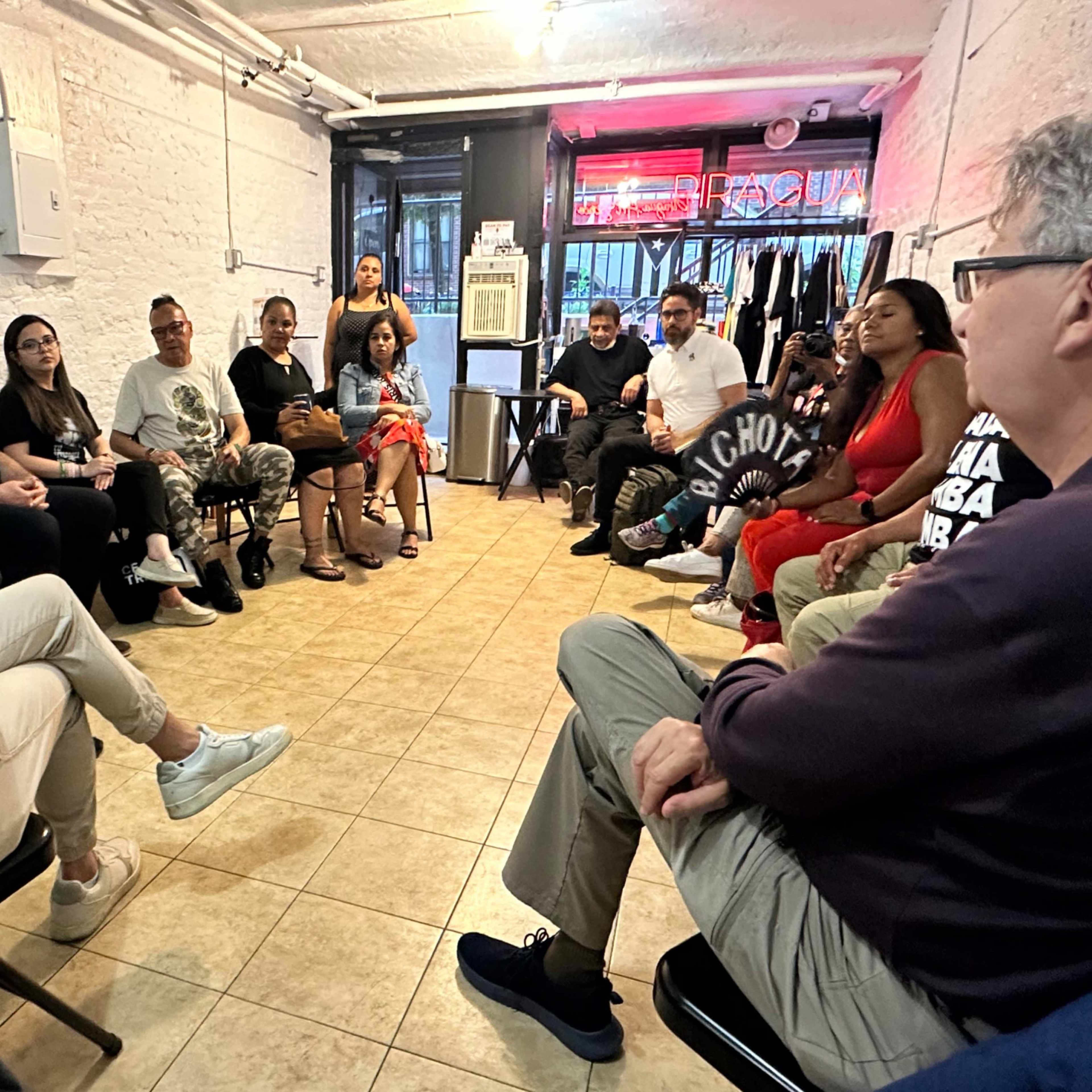 A group of individuals sits in a semi-circle during a gathering in a room with exposed brick walls and clothing displayed in the background.