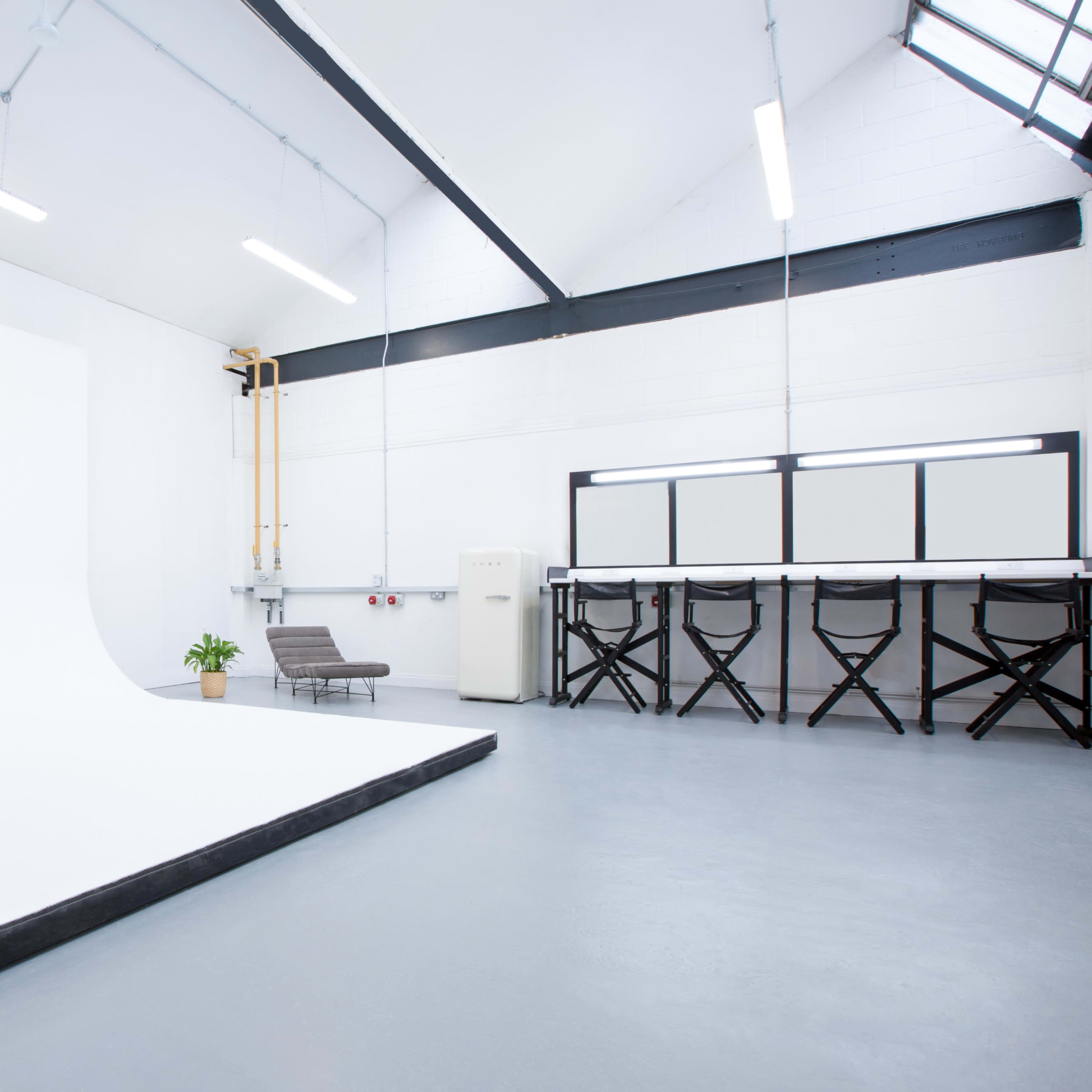 A spacious photography studio featuring a white backdrop, a small potted plant, a gray couch, and a counter with high chairs.