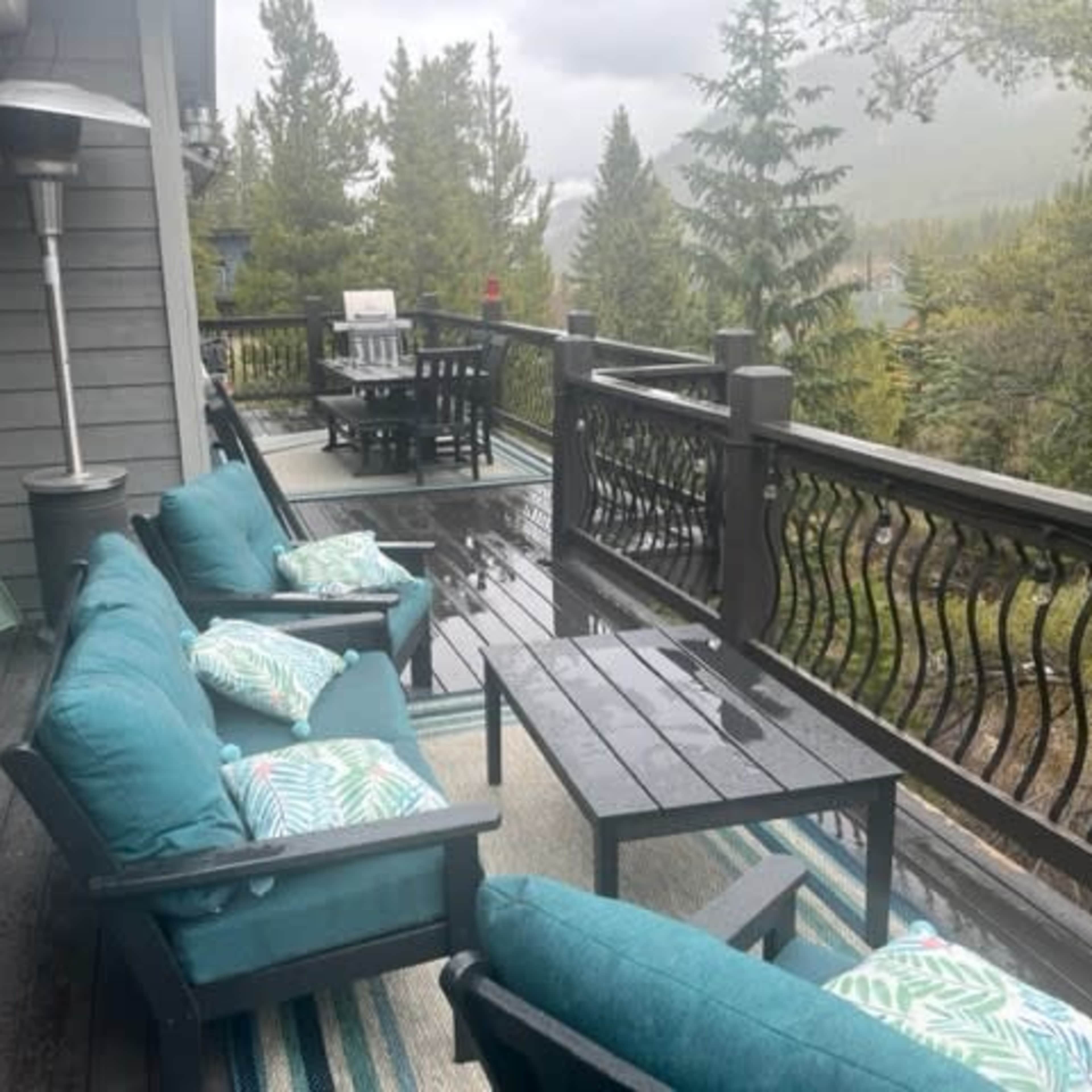 A covered outdoor patio features turquoise seating, a table, and dining furniture, surrounded by tall trees and a mountain backdrop.