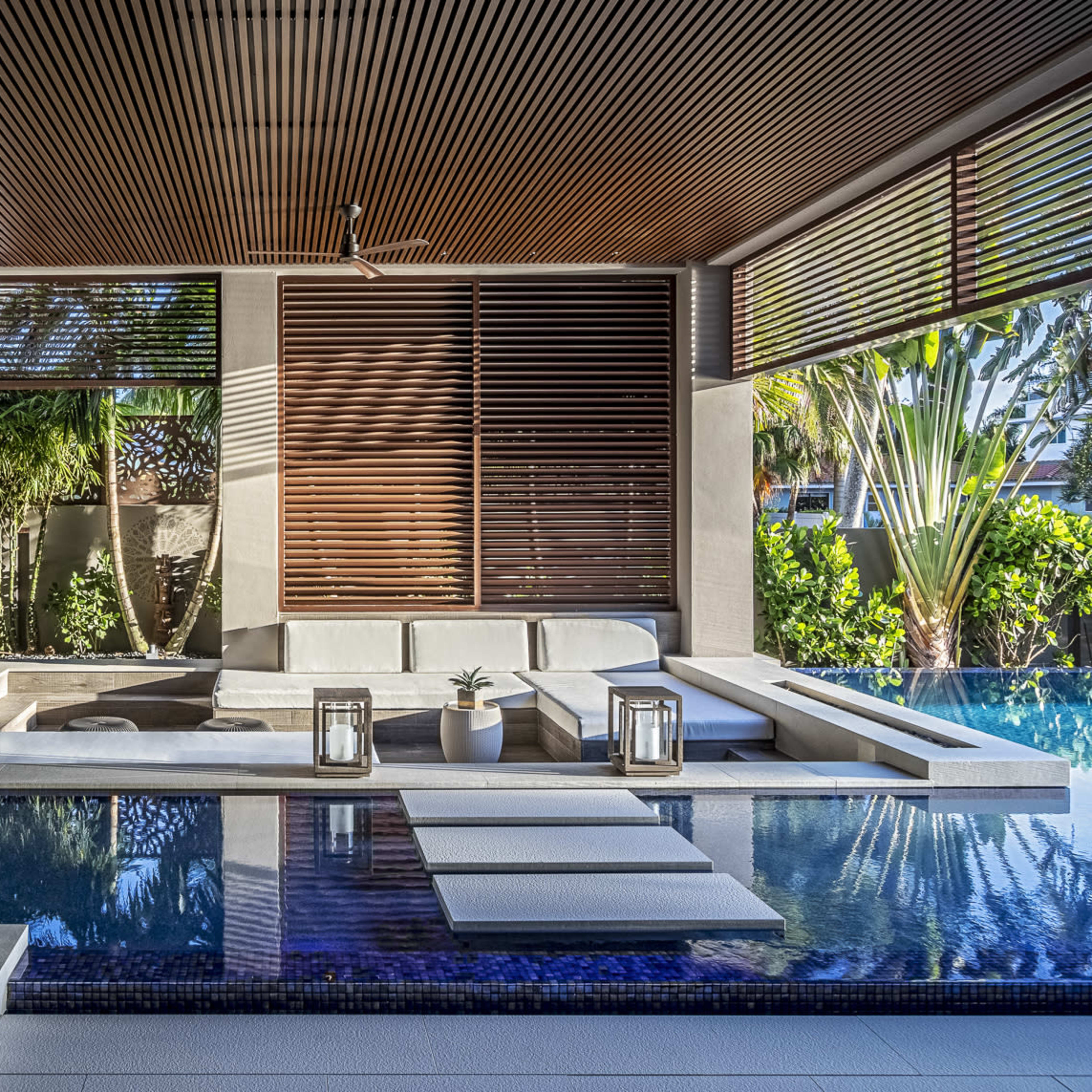 A modern living space featuring a pool area with a sofa, lanterns, and tropical greenery visible through large windows.