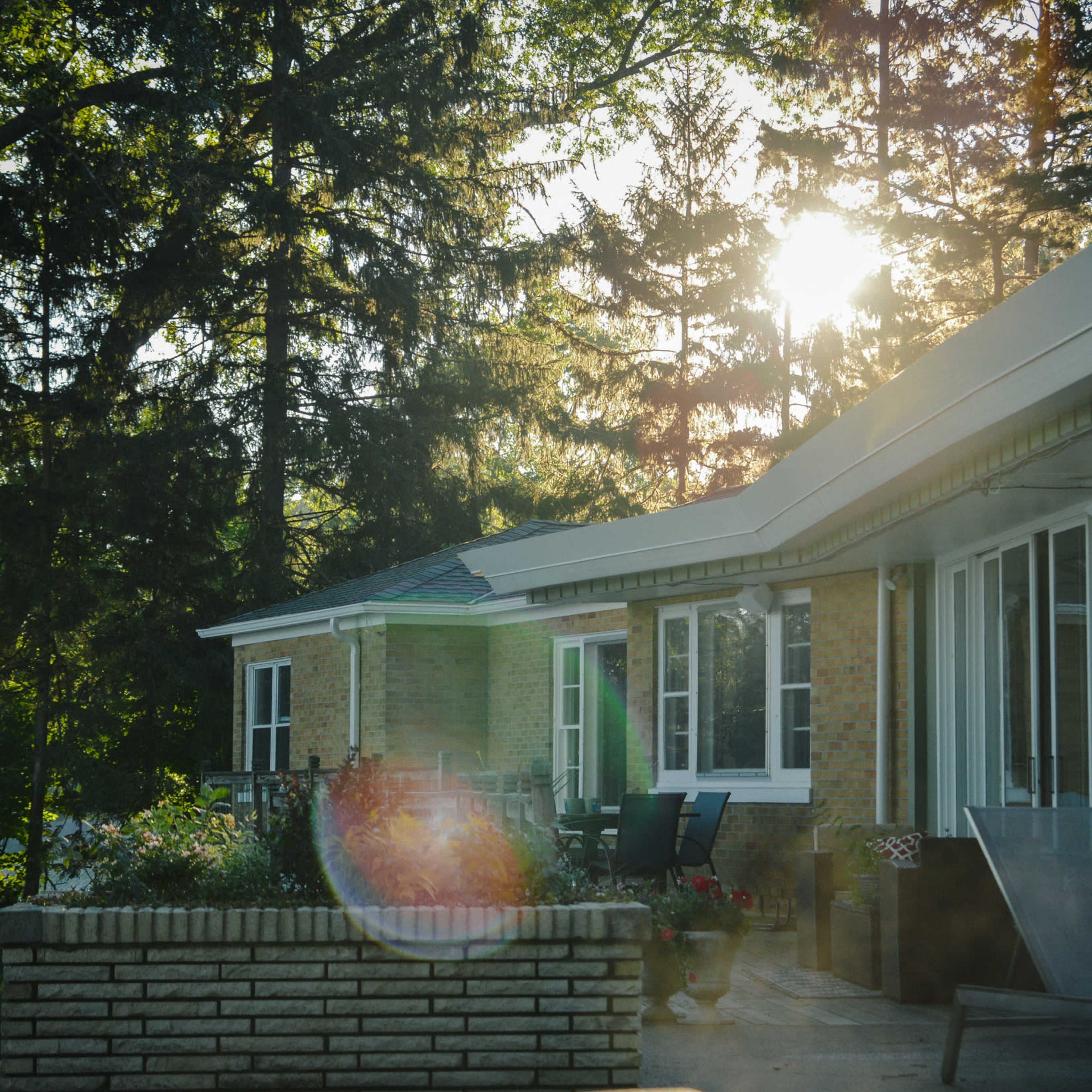 A brick house with large windows is surrounded by trees, with sunlight filtering through the foliage.