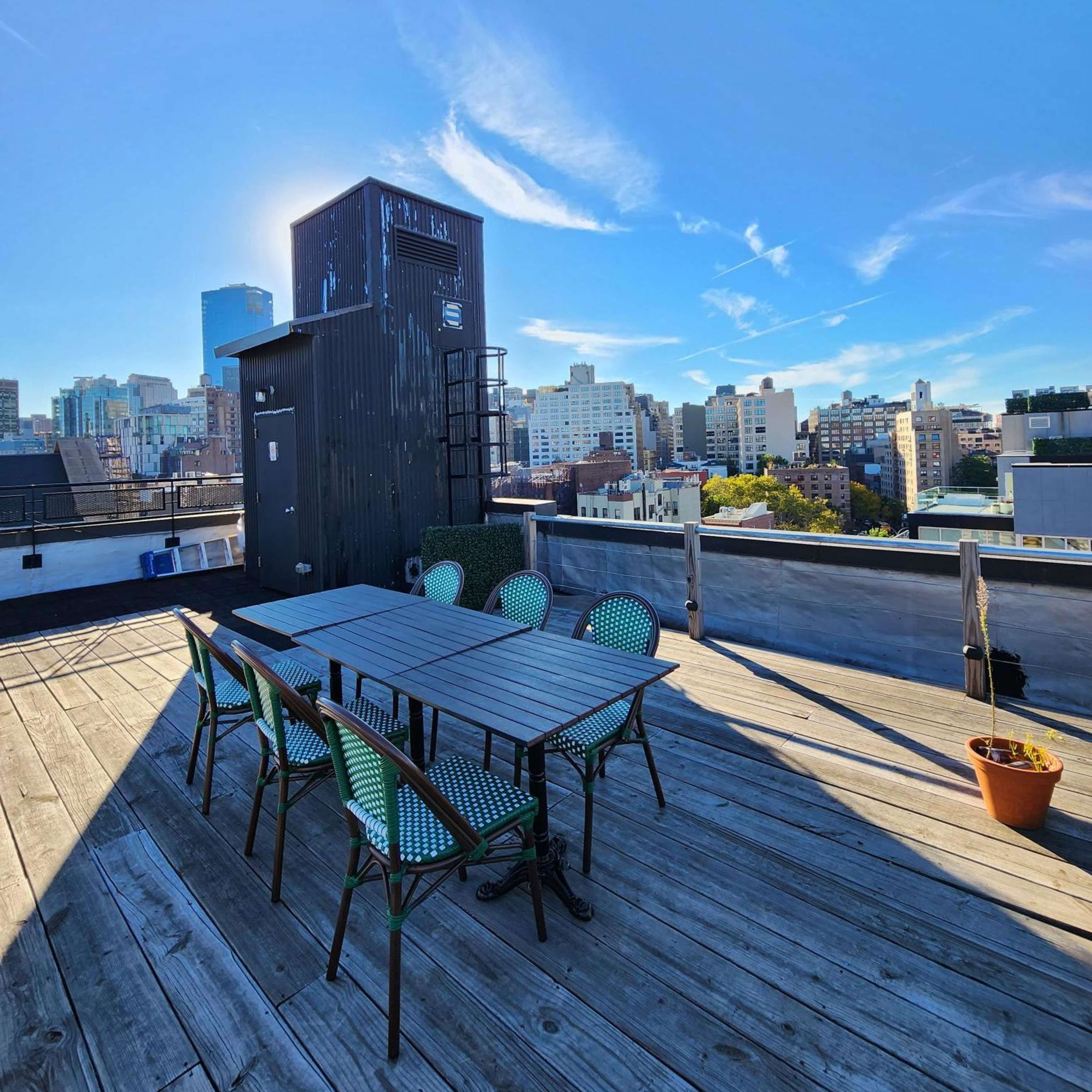 A rooftop terrace features a wooden floor with a metal table and six chairs, overlooking a city skyline under a clear blue sky.