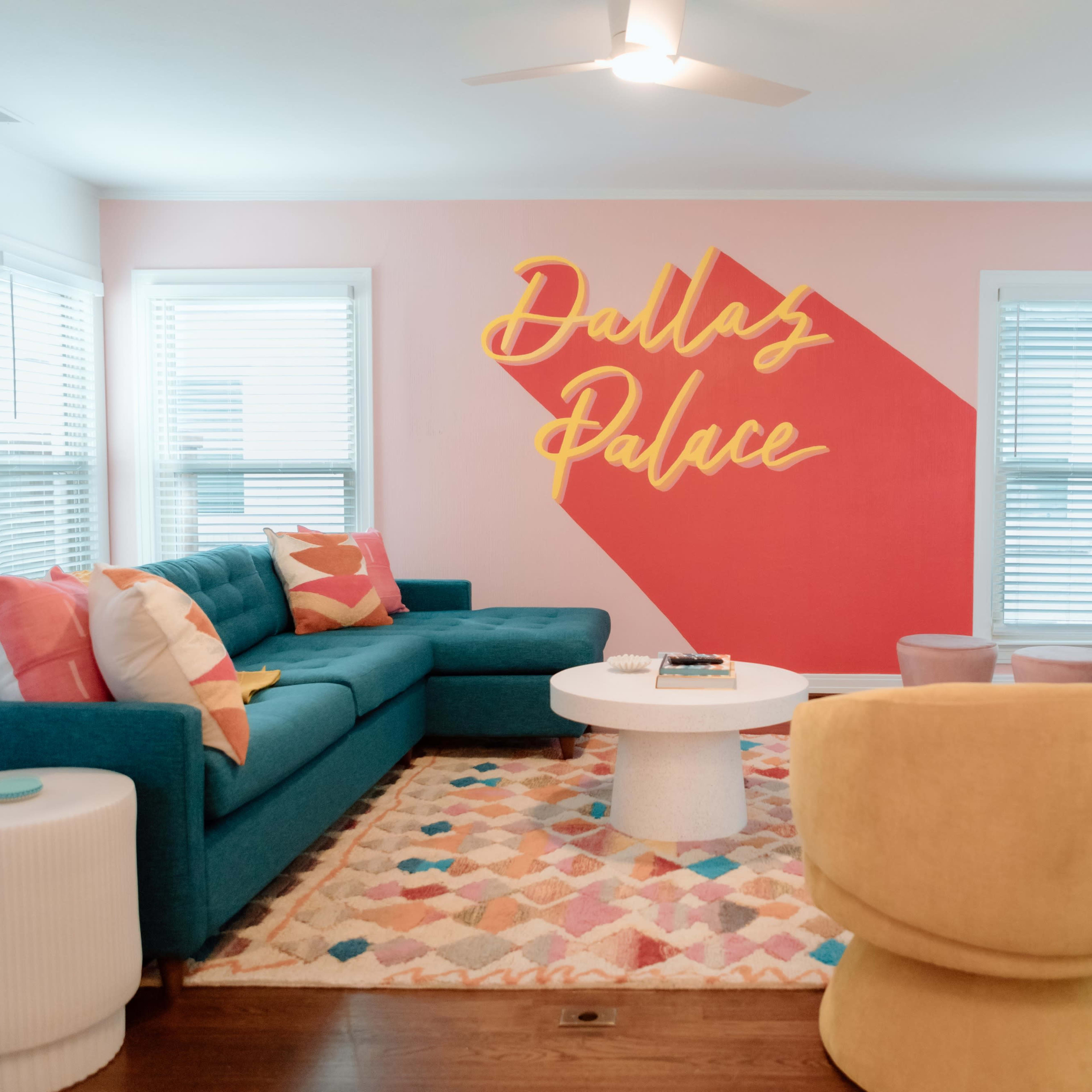 A living room features a turquoise sectional sofa, a round white coffee table, a colorful area rug, and a wall with the text "Dallas Palace" in yellow.