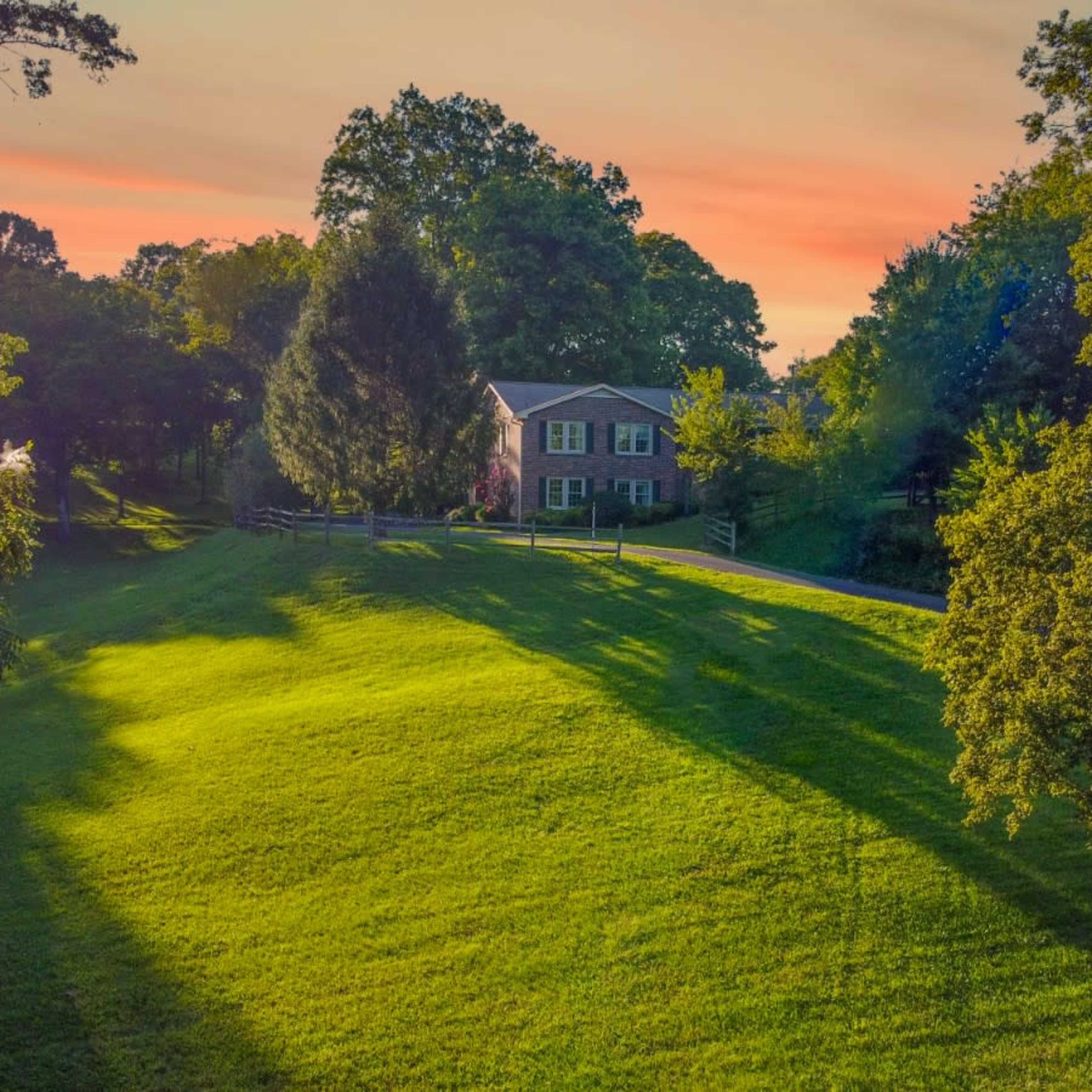 A two-story brick house is situated on a gently sloping green lawn under a colorful sunset sky.