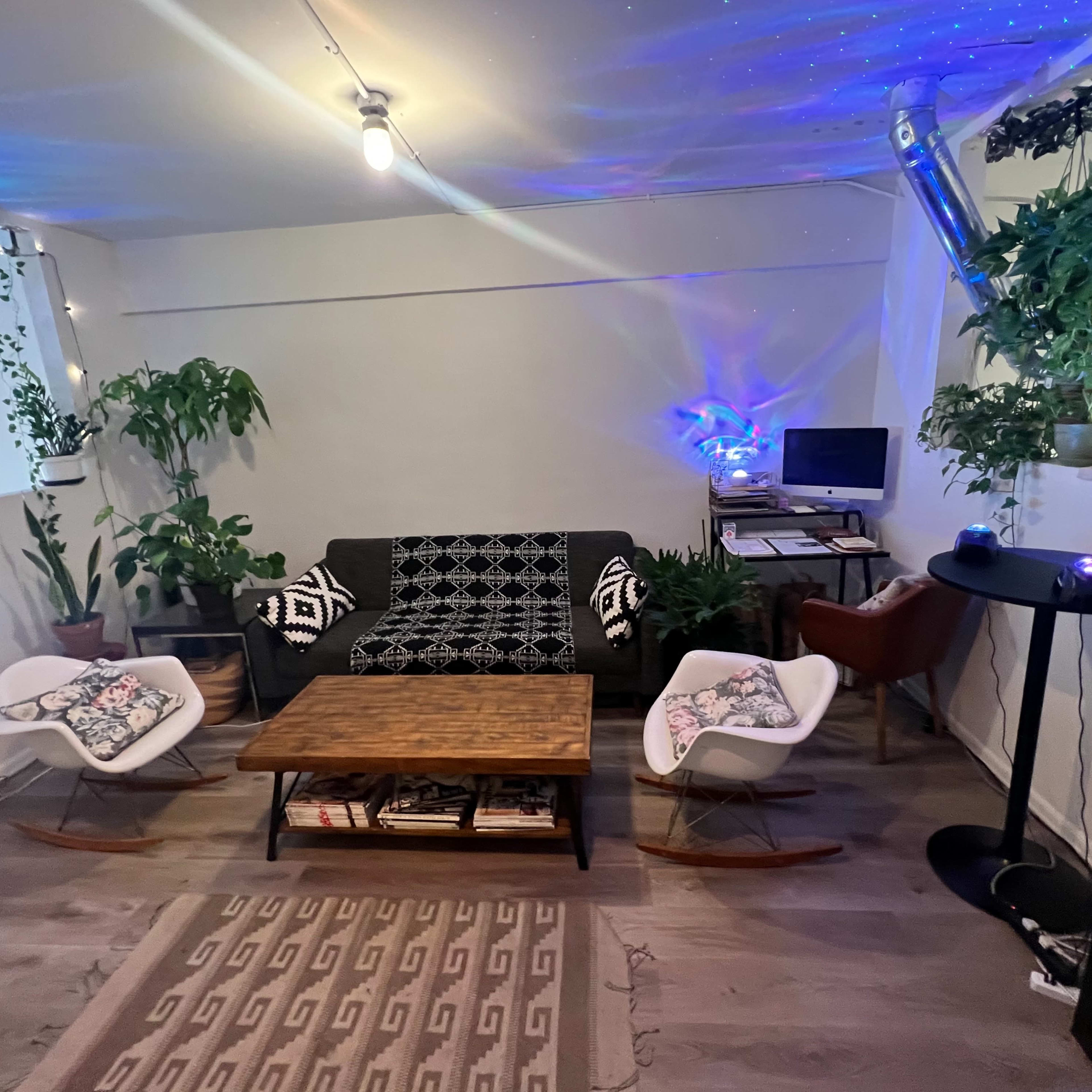 A cozy living room with a black and white patterned couch, two white rocking chairs, a wooden coffee table, and various indoor plants, along with a small desk in the corner.