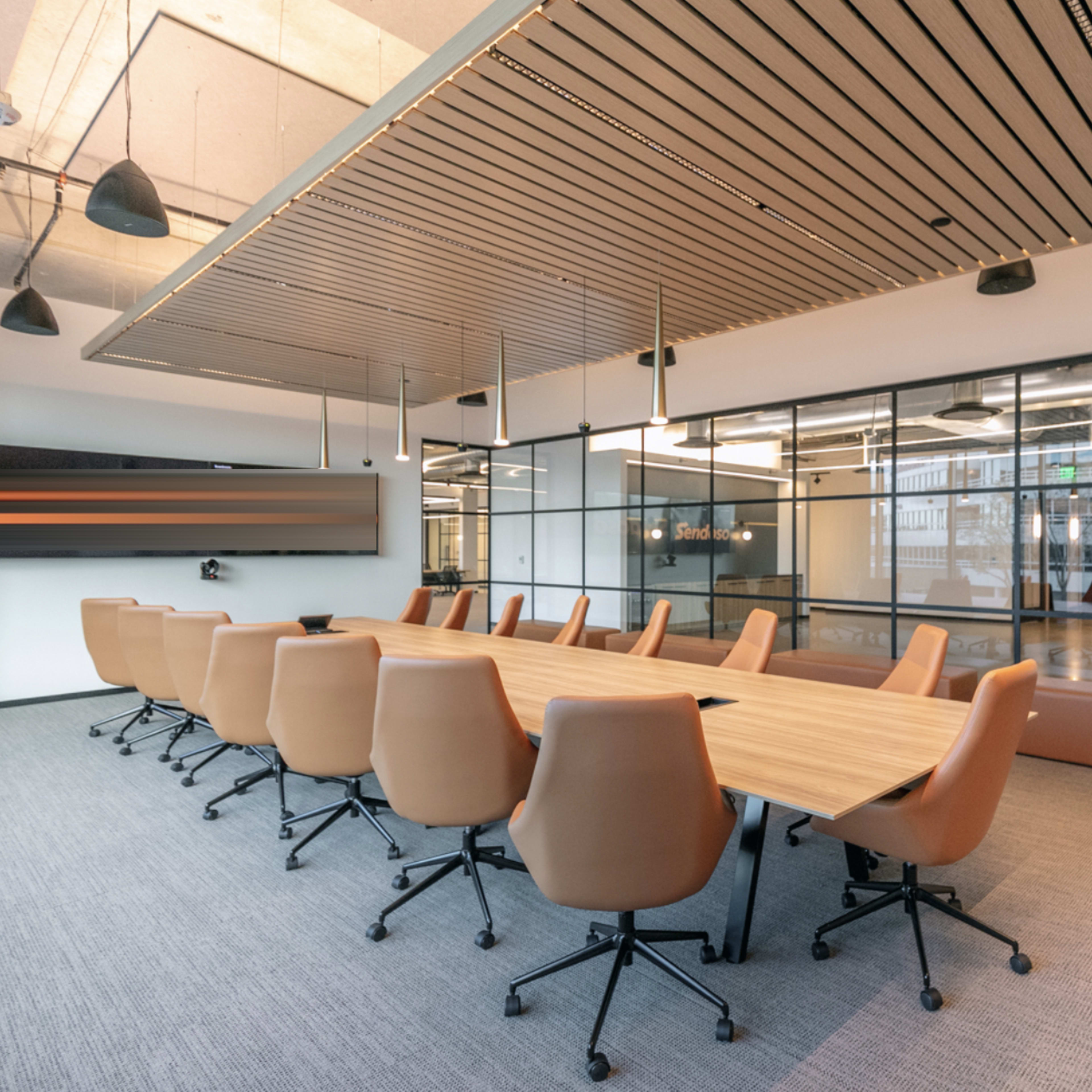 A modern conference room with a long wooden table surrounded by ergonomic chairs and a large screen mounted on the wall.
