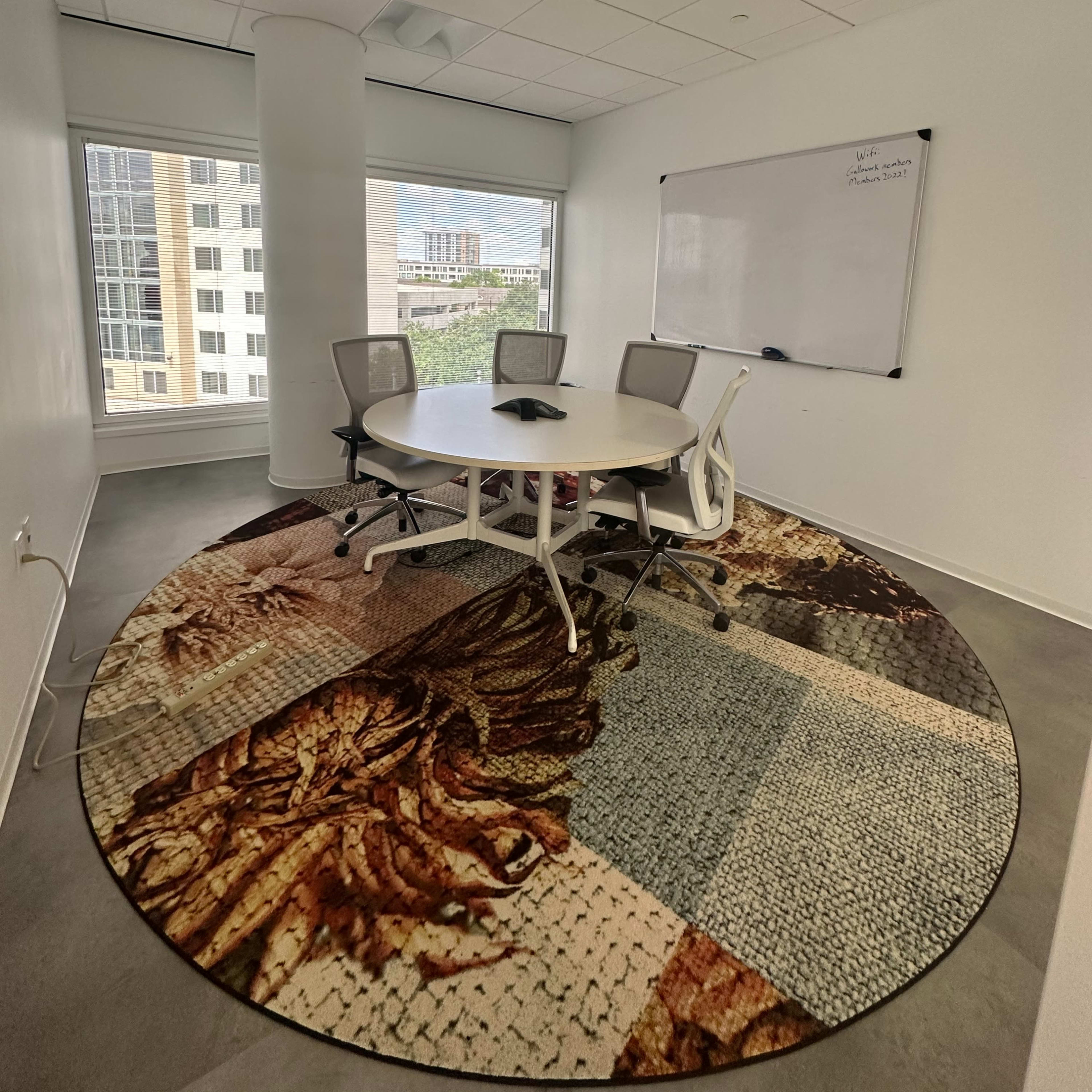 A conference room features a round rug with a detailed design, a whiteboard, a round table, and four chairs, with large windows providing natural light.