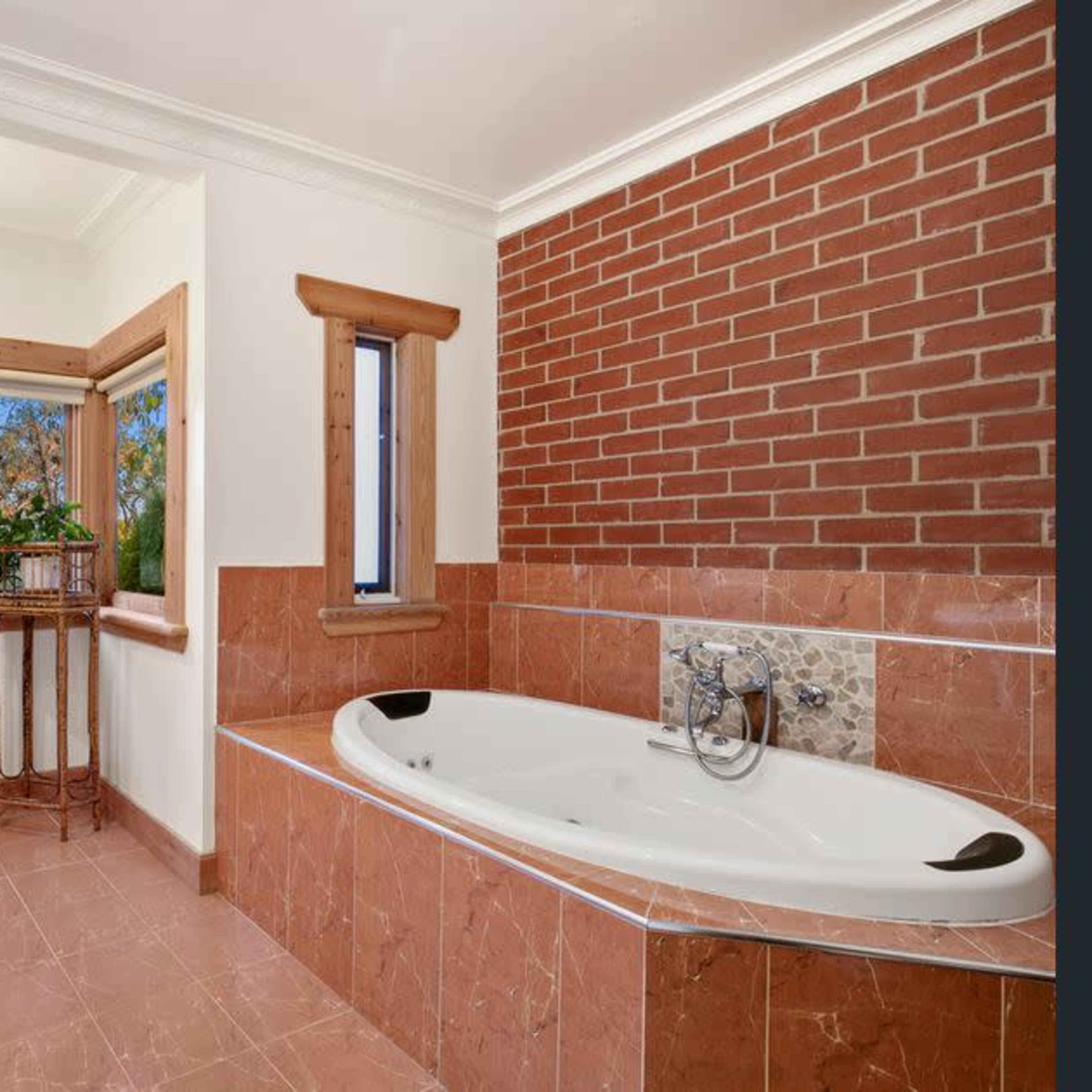 A spacious bathroom featuring a corner bathtub, a vintage sink, and a window with a view of greenery surrounded by brick and tiled walls.