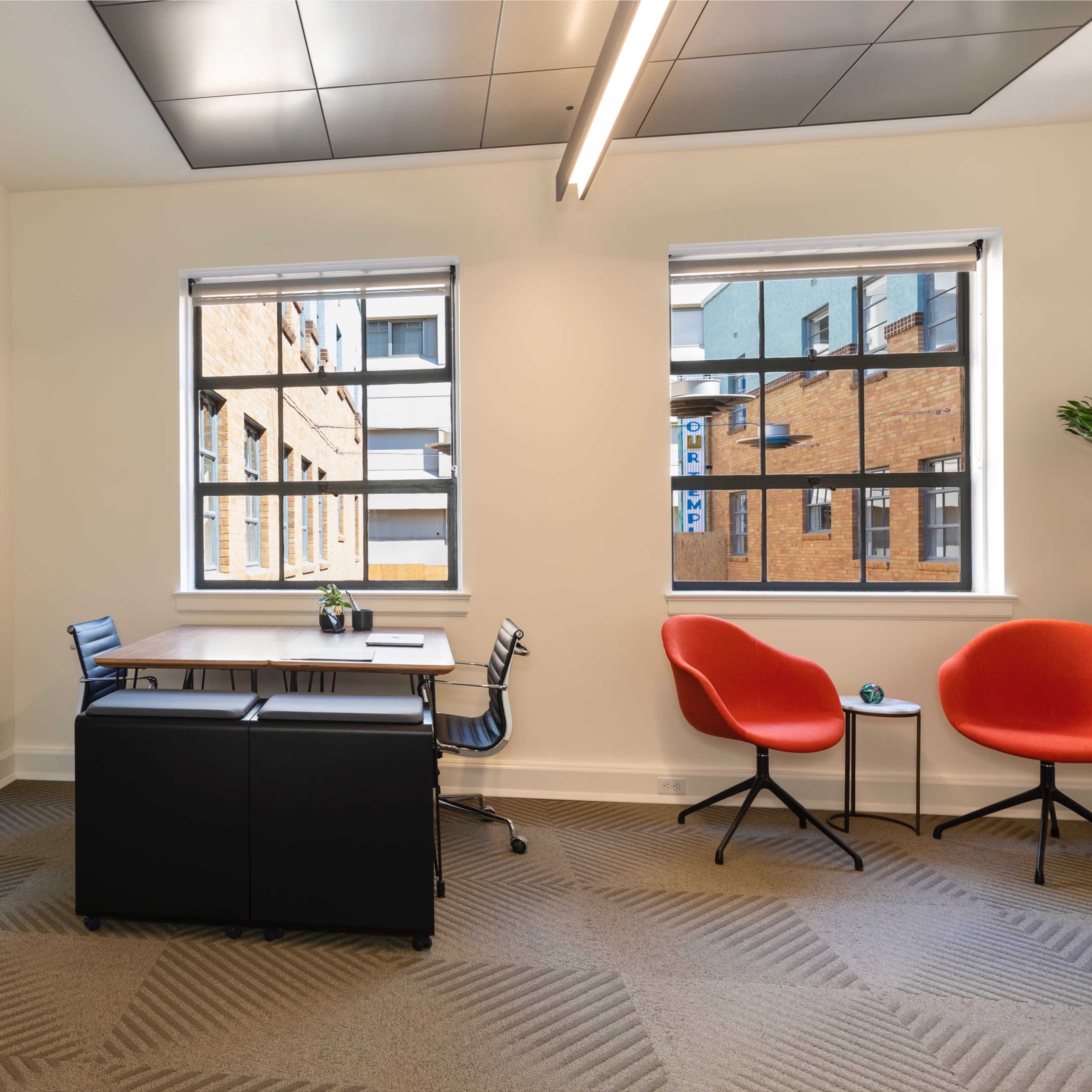 A modern office space with a desk and chair, two red chairs, a shelving unit, and large windows allowing natural light to enter.