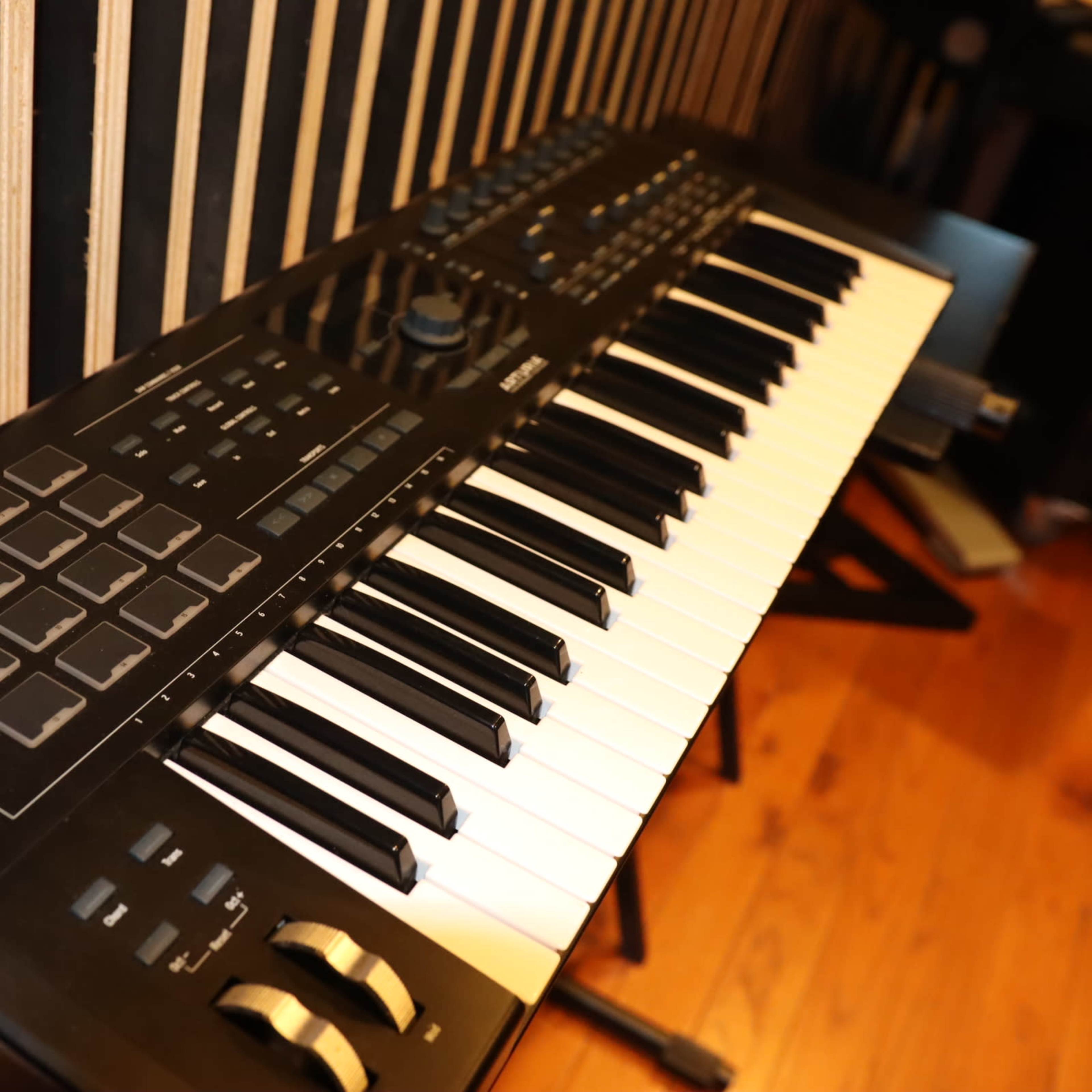 A black MIDI keyboard with white keys and control pads rests on a wooden floor next to striped walls.