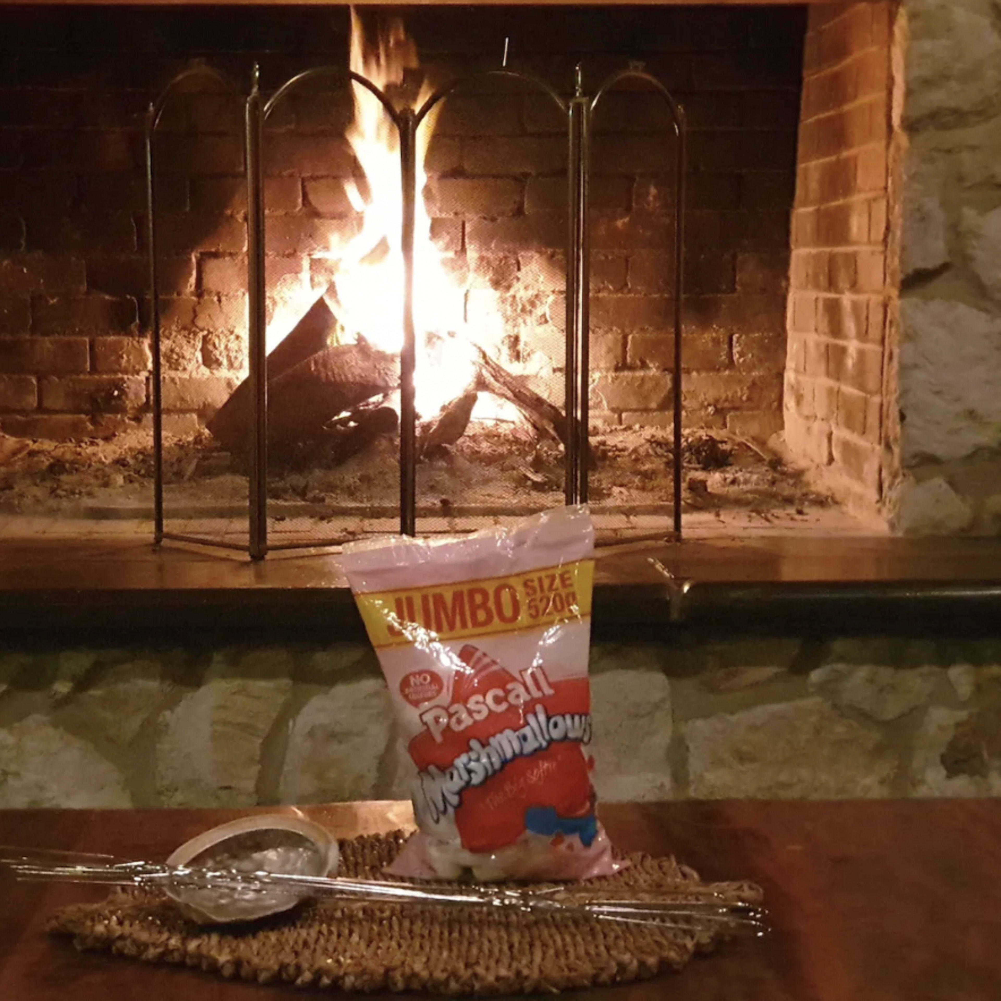 A bag of jumbo marshmallows is placed on a table in front of a stone fireplace with flames burning.