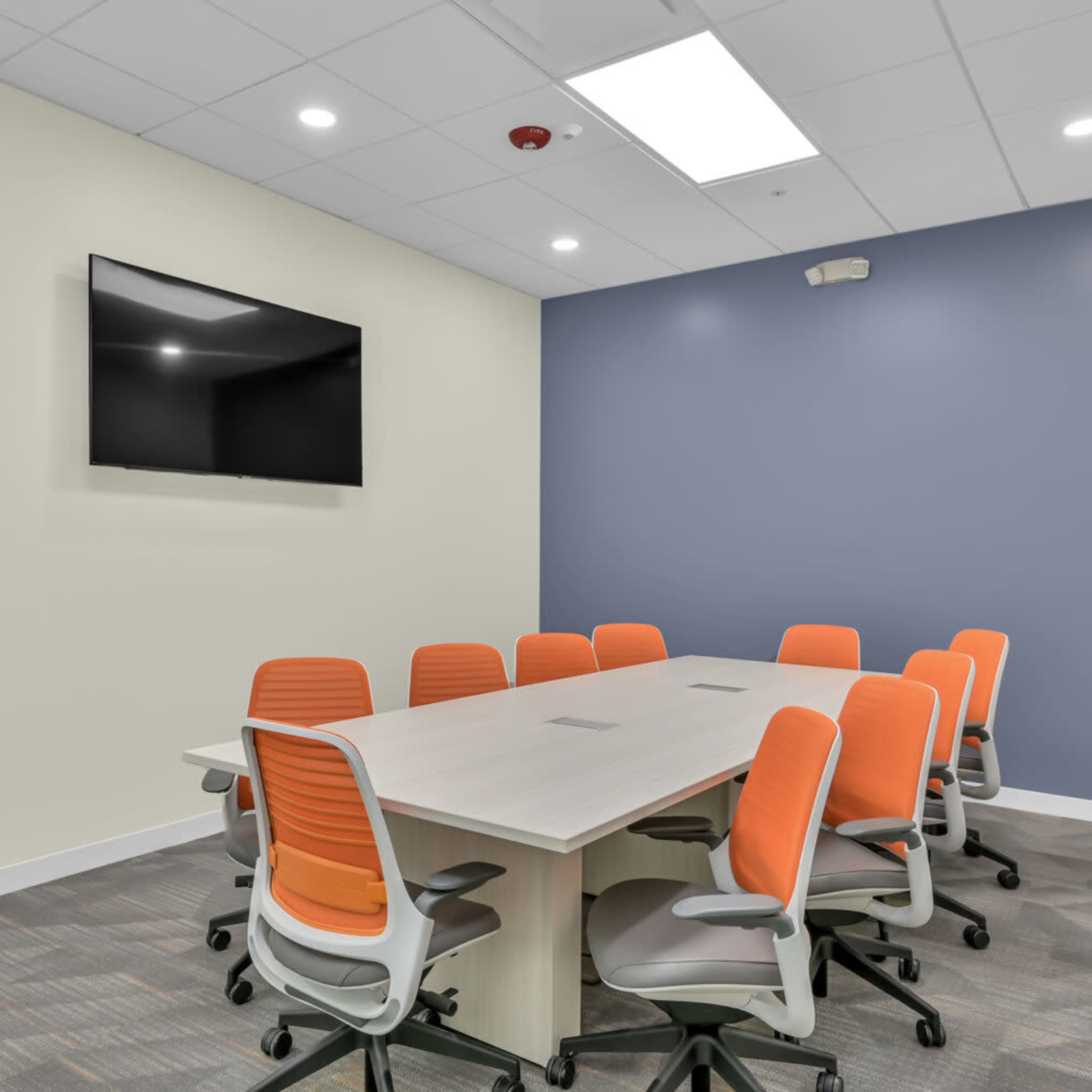 A modern conference room features a large table surrounded by eight orange and gray chairs, with a wall-mounted television and light-colored walls.